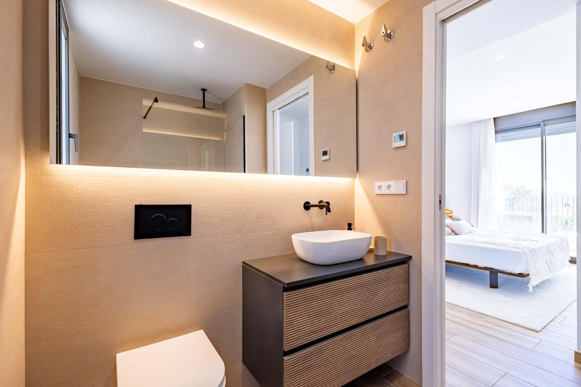 Modern bathroom with a white vessel sink on a dark wood vanity and a large backlit mirror; doorway opens to a bright bedroom.