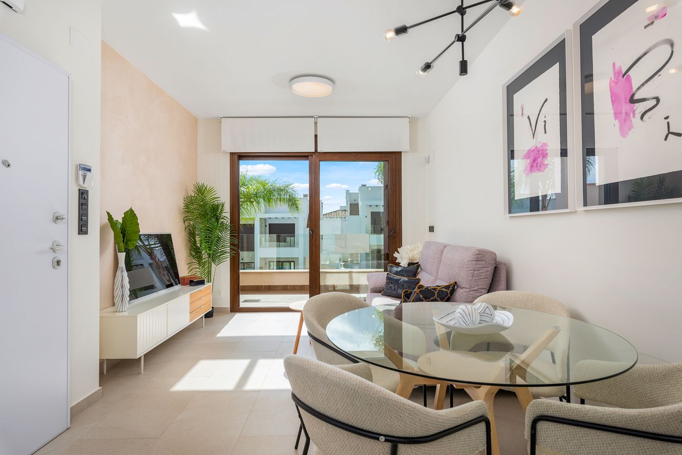 Bright living-dining area with a round glass table, beige chairs, and a pink sofa near a large glass sliding door to a balcony outside.