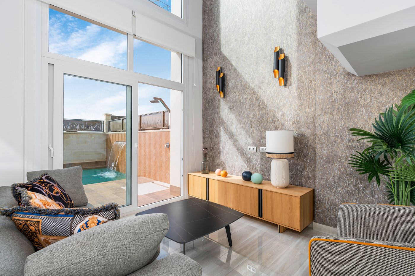 Modern living room with large glass doors to a pool, gray sofa with patterned cushions, and a wood sideboard in front of a textured wall.