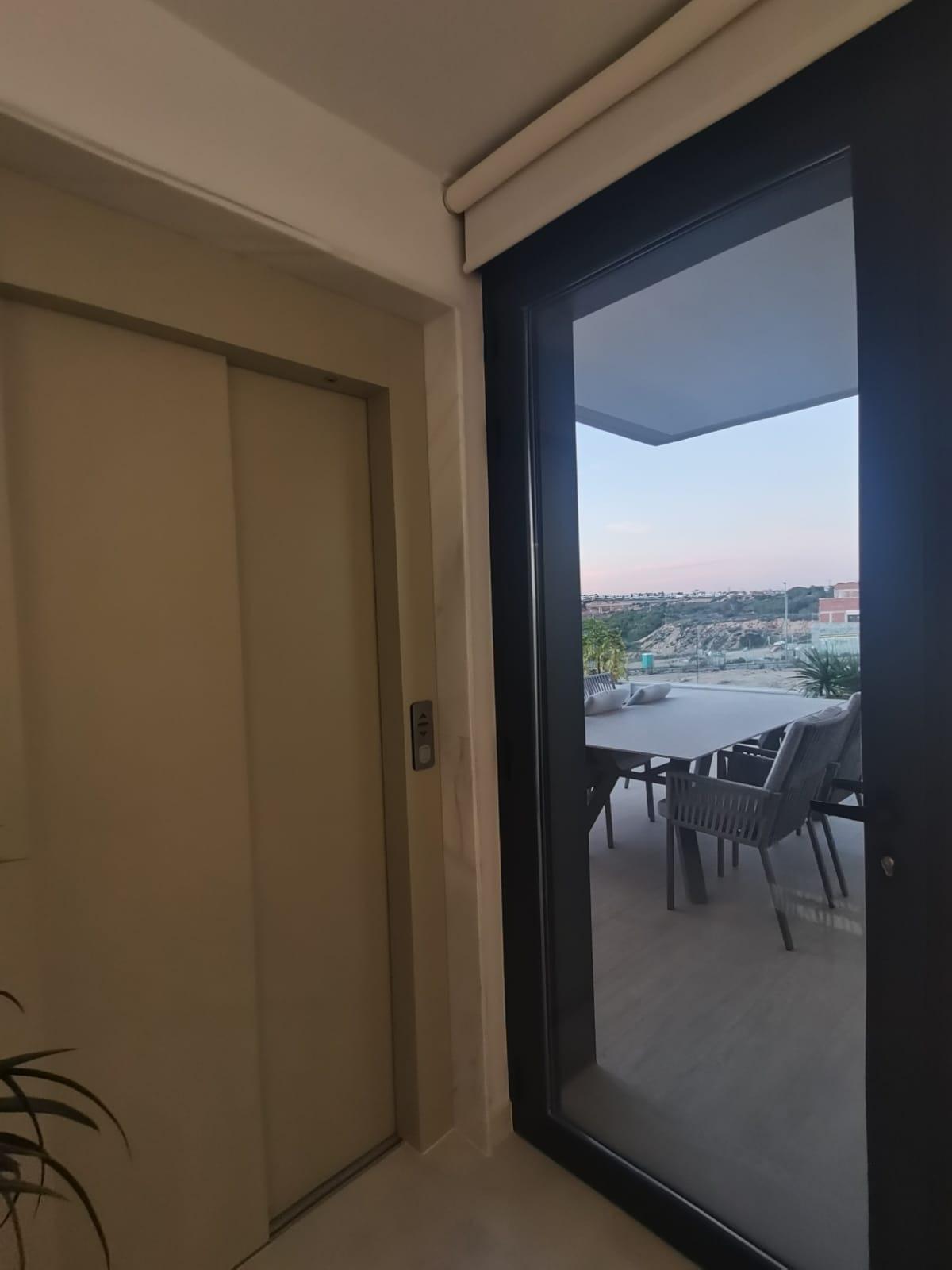 Indoor hallway with a closed elevator door on the left and a glass balcony door opening to an outdoor dining area with a table and chairs.