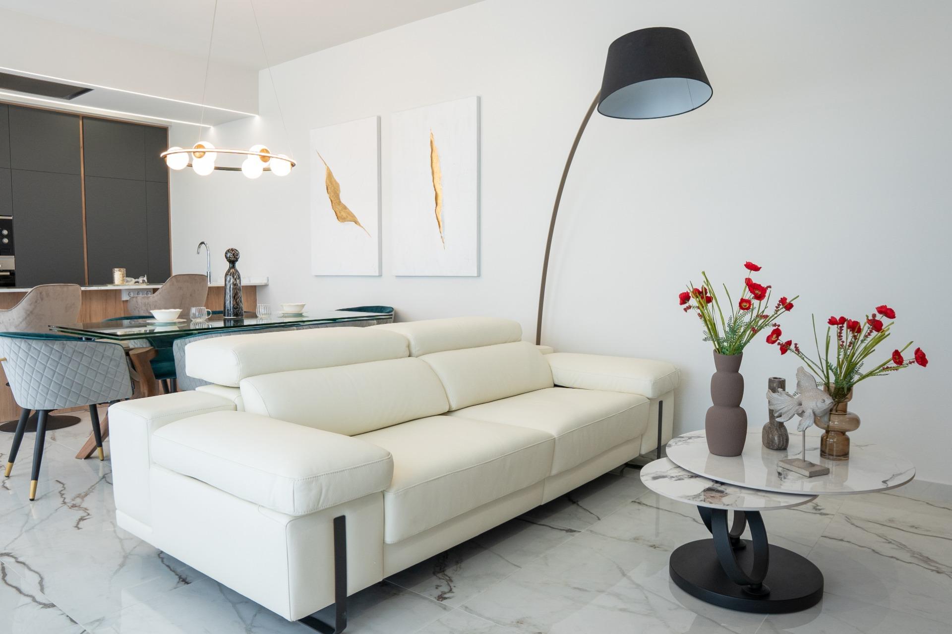 Bright modern living room with a white leather sectional, glass dining table, and a large black curved floor lamp against a white wall; red flowers in a vase on a marble coffee table.
