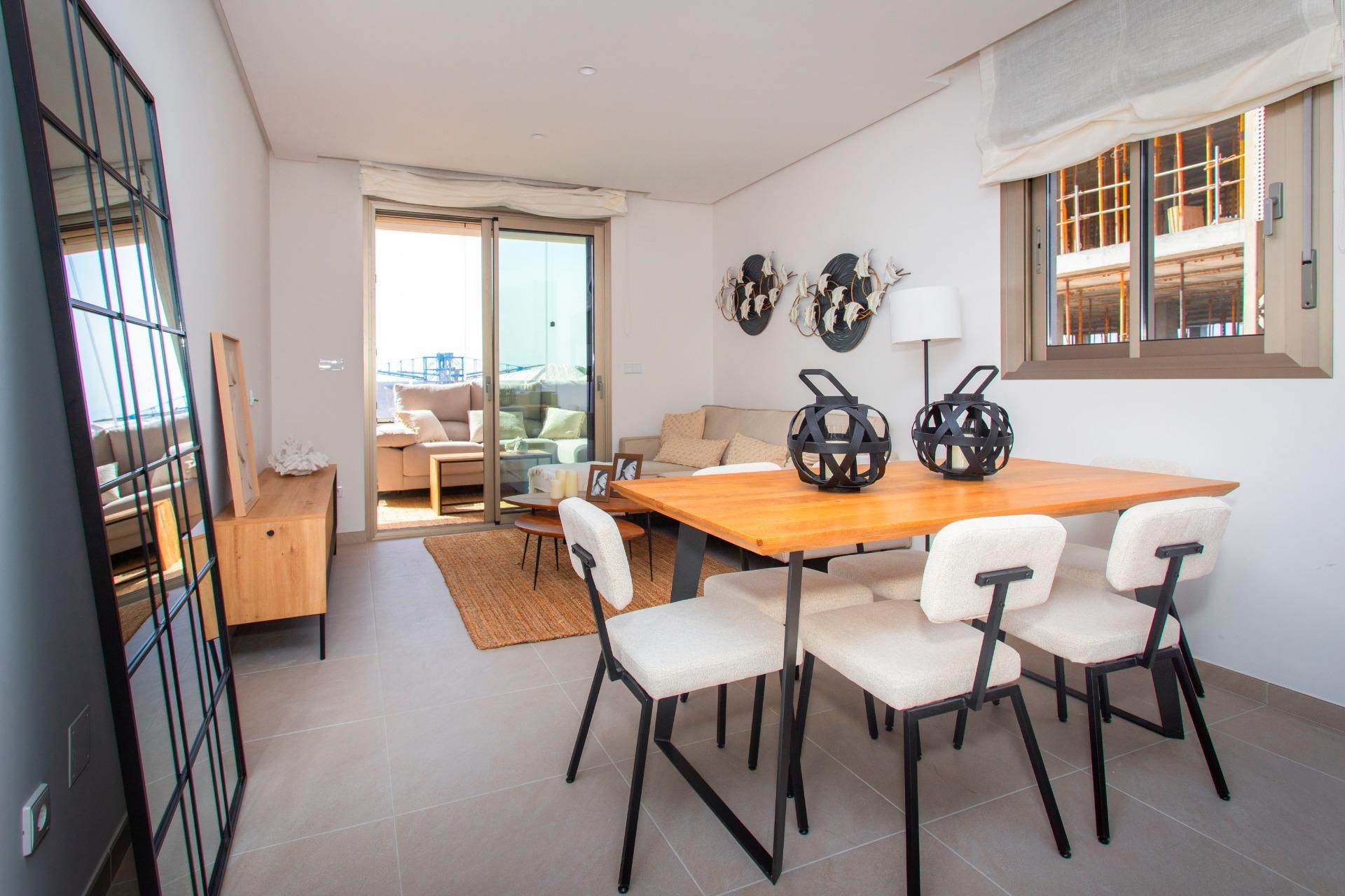 Open-plan dining area with a wooden table and white upholstered chairs, adjacent living area and sliding glass door to a balcony.