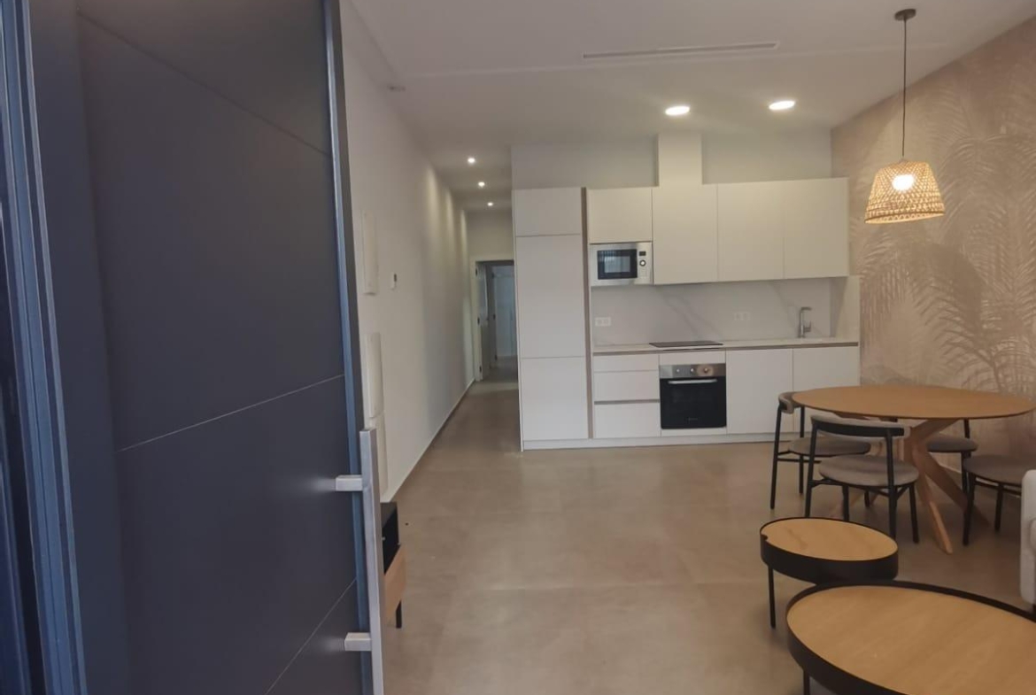 Open doorway into a modern kitchen: white cabinets, built-in oven and microwave, and a round wooden dining table with chairs nearby.
