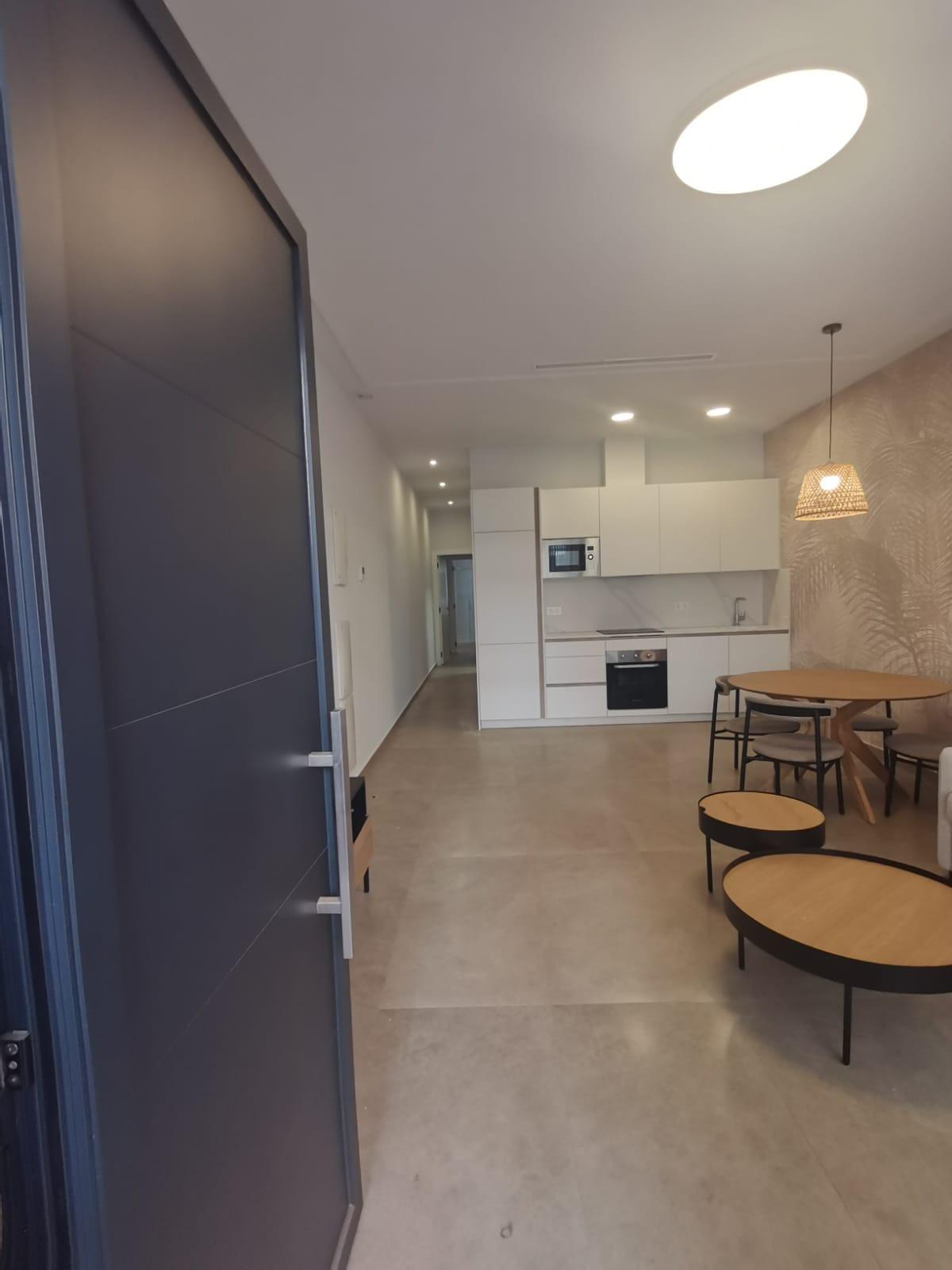 Open doorway into a modern kitchen: white cabinets, built-in oven and microwave, and a round wooden dining table with chairs nearby.