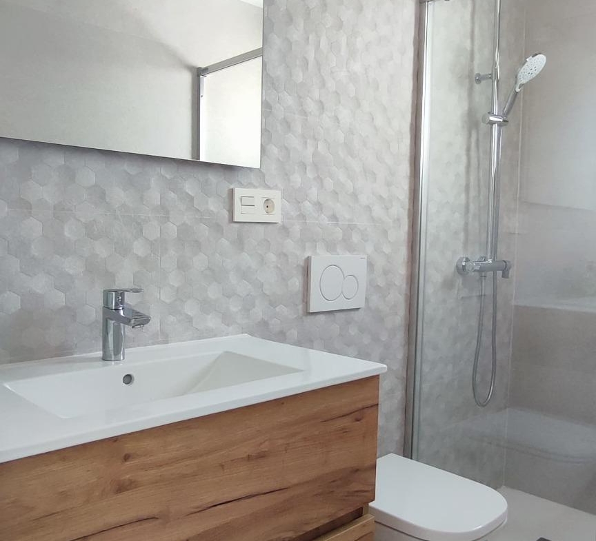 Modern bathroom with a wooden vanity, white sink, and wall-mounted faucet, a large mirror, gray hex-patterned walls, and a glass shower enclosure with a handheld showerhead.