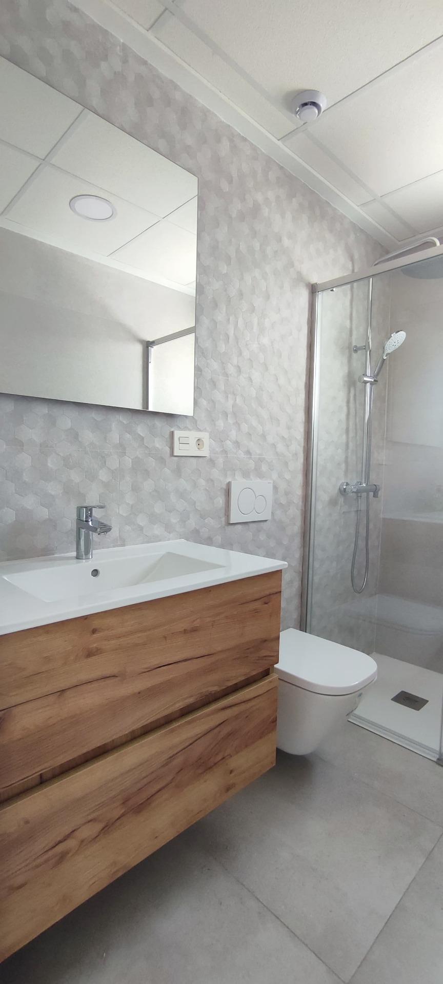 Modern bathroom with a wooden vanity, white sink, and wall-mounted faucet, a large mirror, gray hex-patterned walls, and a glass shower enclosure with a handheld showerhead.