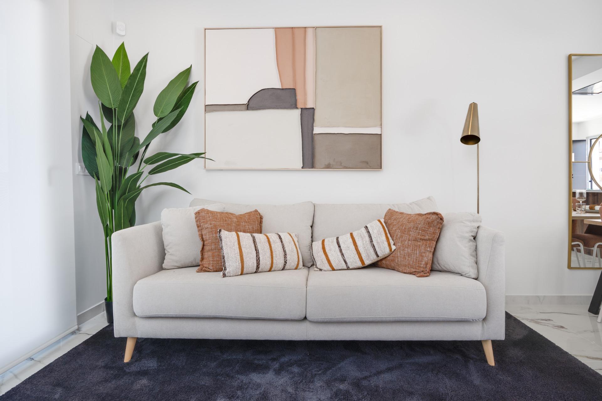 Bright living room with a light gray sofa, orange striped cushions, and a large beige abstract painting above; tall green plant to the left.