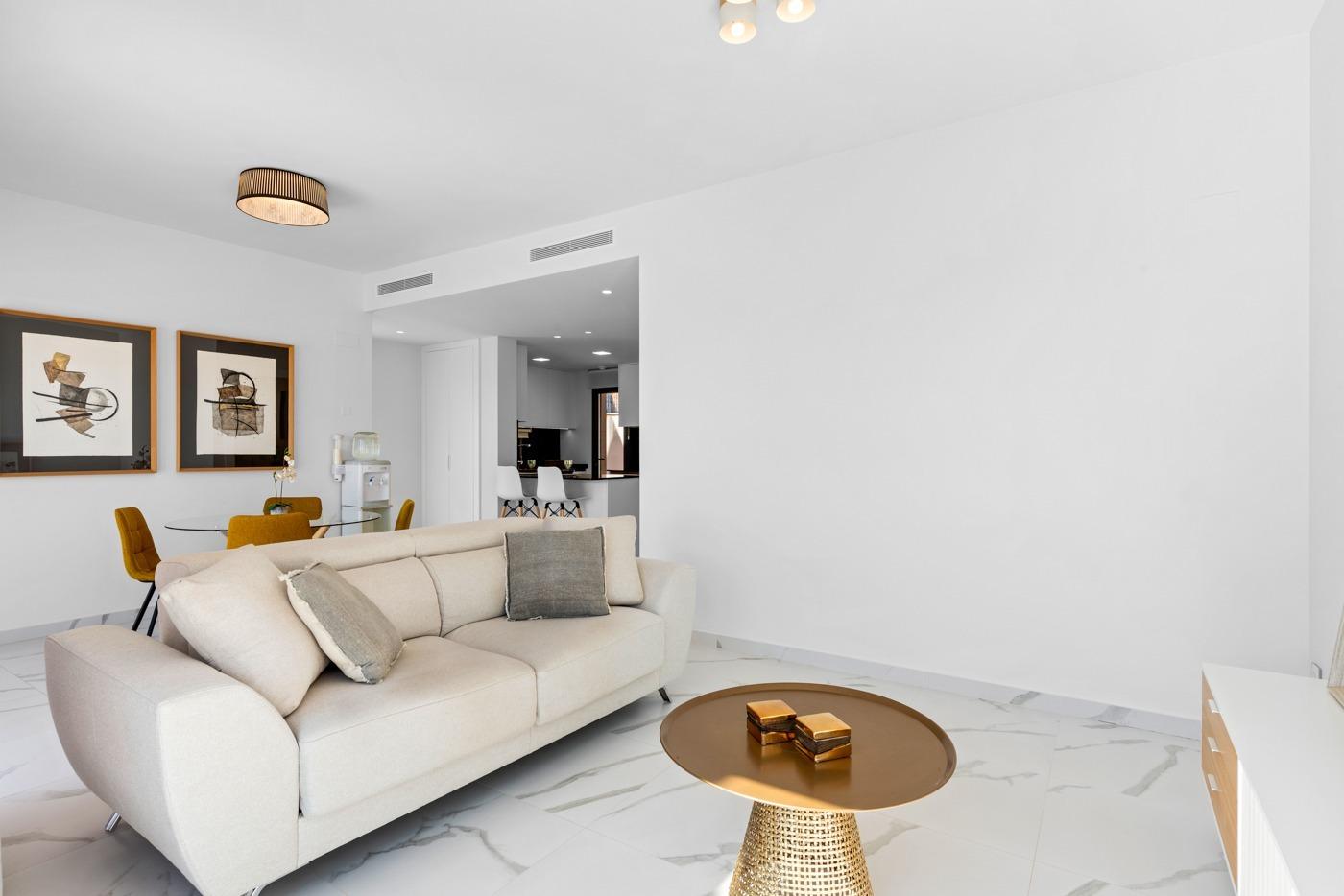 Bright, minimalist living room with a beige sofa, marble floor, and a round gold coffee table; open-plan kitchen in the background.