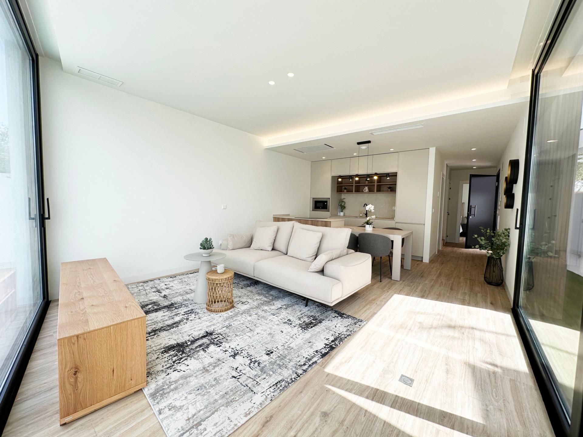 Open-plan living area with a pale beige sectional sofa, rug, and a kitchen visible in the background behind glass doors.