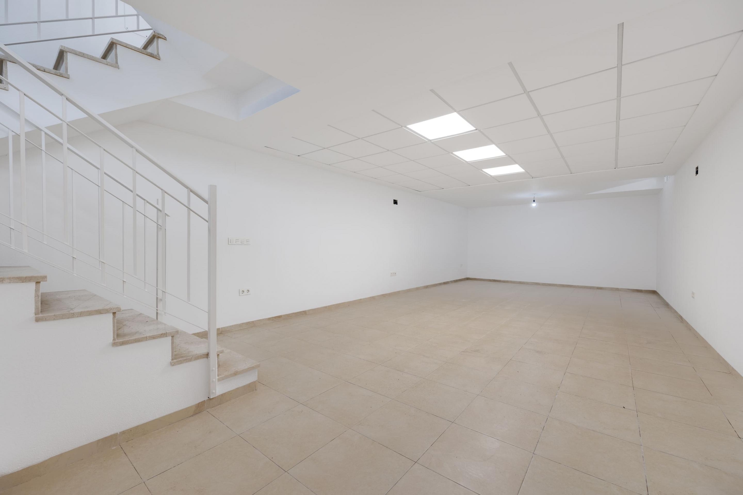 Empty white interior space with a left-side staircase featuring a metal railing, beige tile floor, and a grid ceiling with lights.