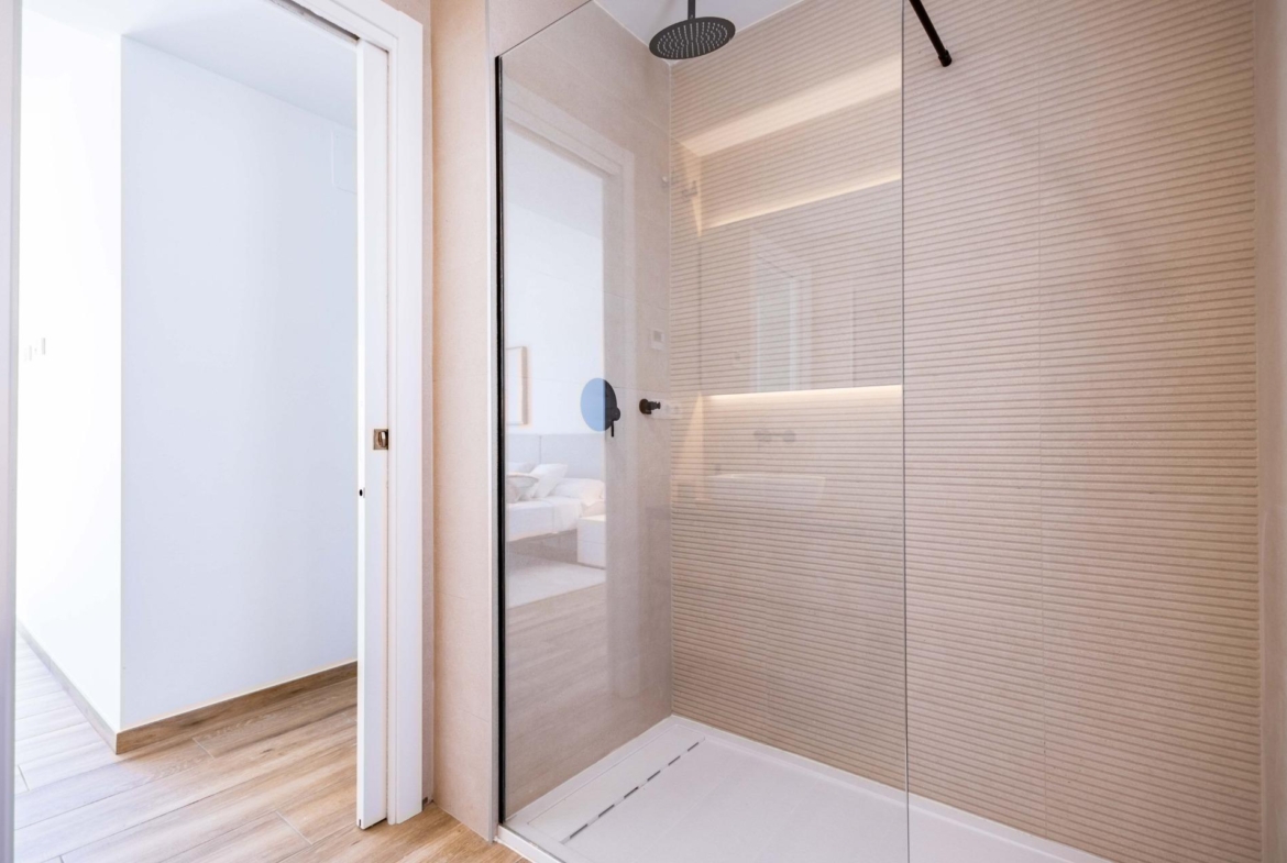 Modern glass-walled shower with a large round rain shower head and beige textured tile walls in a contemporary bathroom.