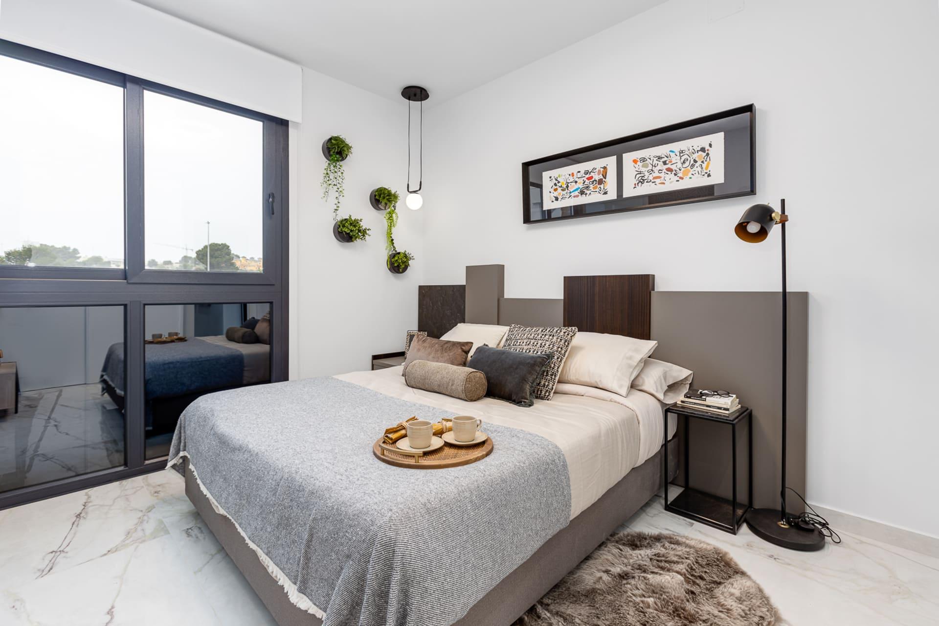 Contemporary bedroom with a gray upholstered bed, matching bedding, and a tray of cups on the quilted blanket. A large glass window and plants decorate the space.