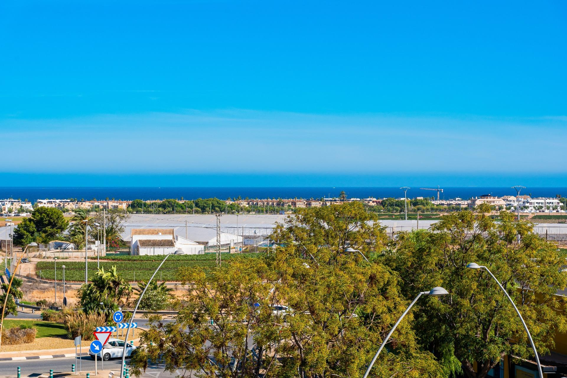 Coastal town seen from a road roundabout in the foreground, with trees and the blue sea on the horizon under a clear sky.