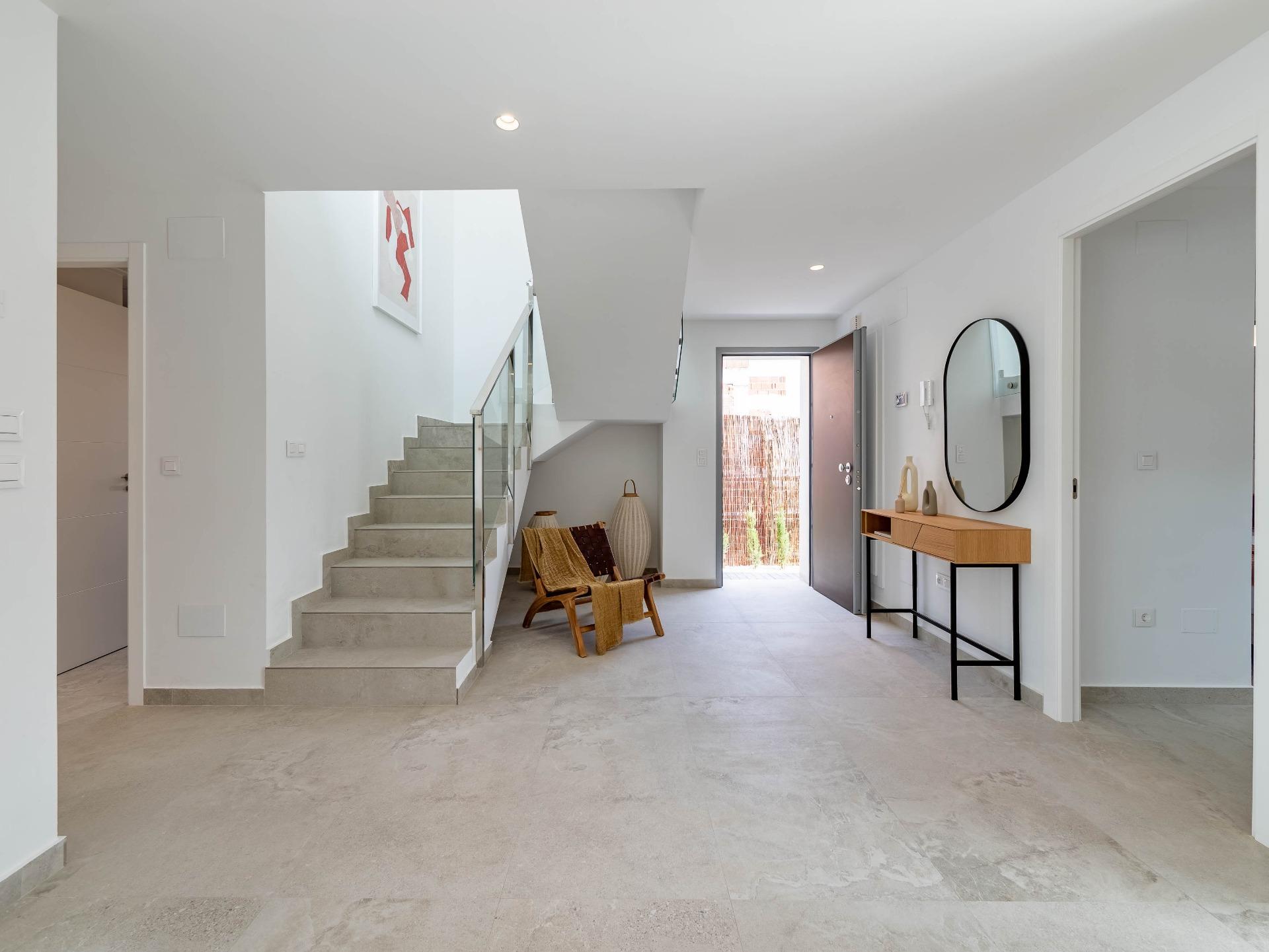 Bright minimalist foyer with staircase on left, front door open to a courtyard, and a round mirror above a wooden console table on the right.