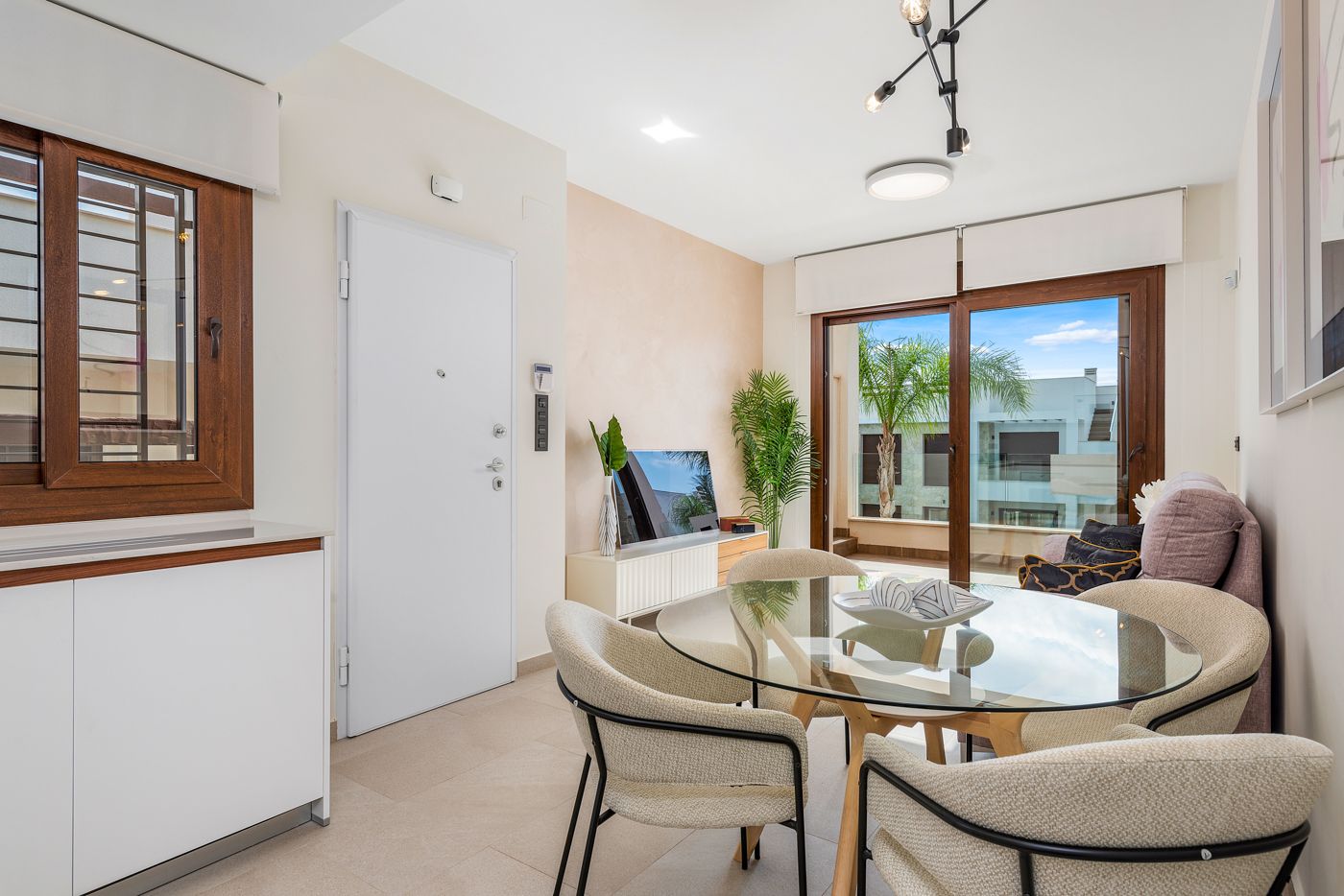 Bright dining/living area with a round glass table, beige chairs, and large sliding doors to a balcony with palm trees outside.