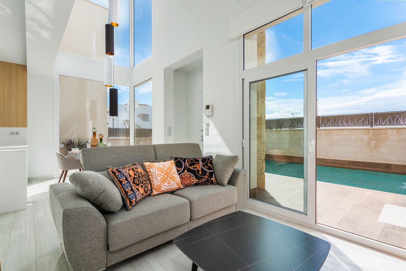 Bright living room with a grey sofa and decorative cushions, large glass doors overlook a pool patio outside provided by floor-to-ceiling windows.