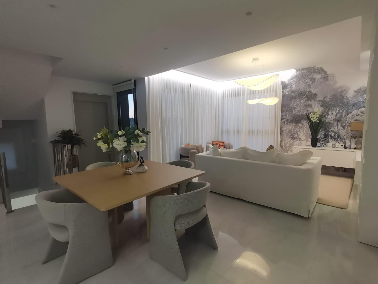 Open-concept living and dining area with a light wood table, gray upholstered chairs, and a white sofa under sheer curtains.