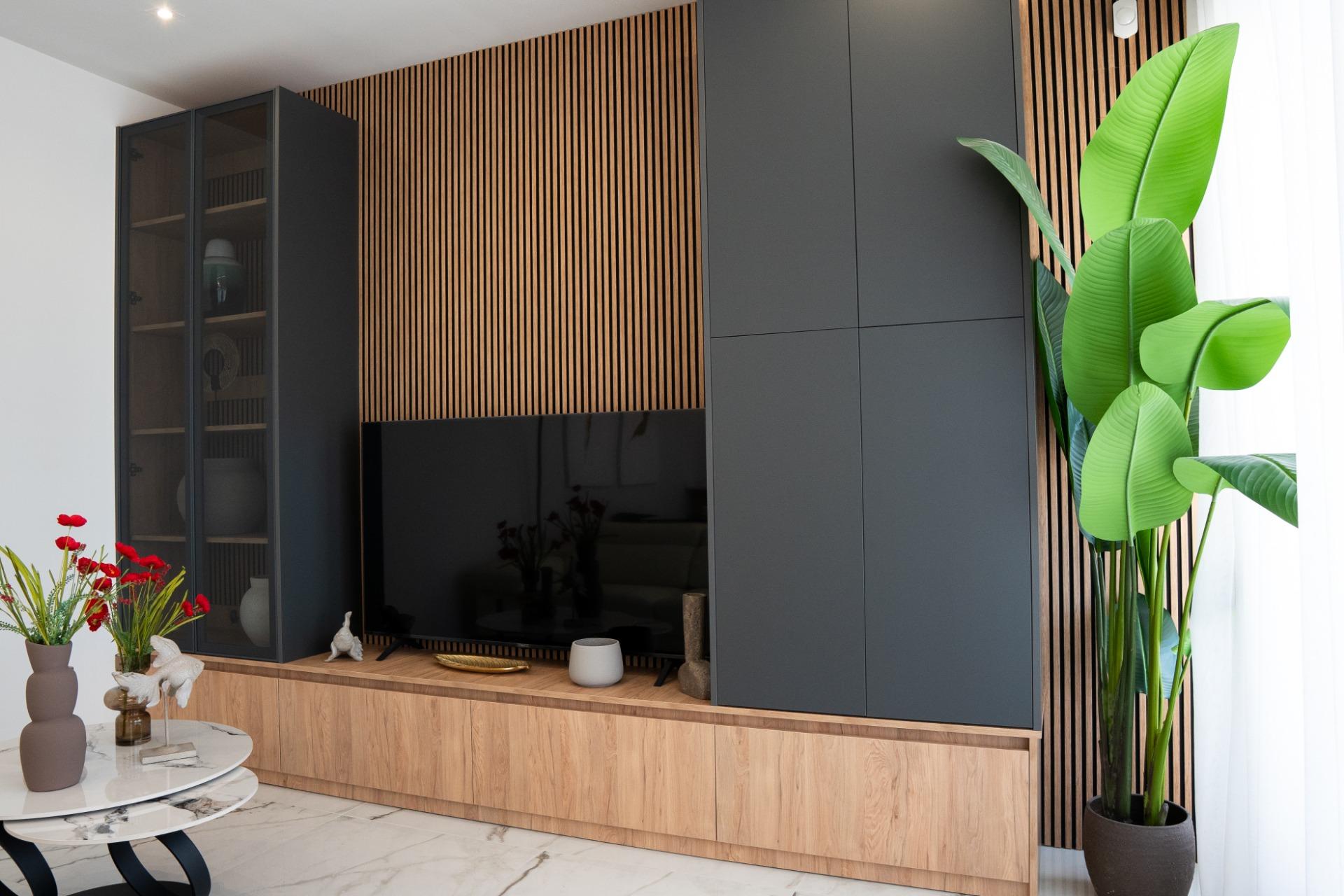 Modern living room cabinet wall with a black tall storage unit, glass-front display cabinet, and a flat-screen TV mounted on a vertical wooden slat accent wall; potted plants nearby.