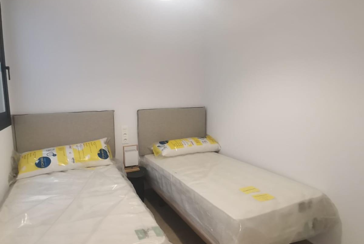 Small twin bedroom with two single beds in plastic, beige headboards, and a shared nightstand beneath a hanging ceiling light.