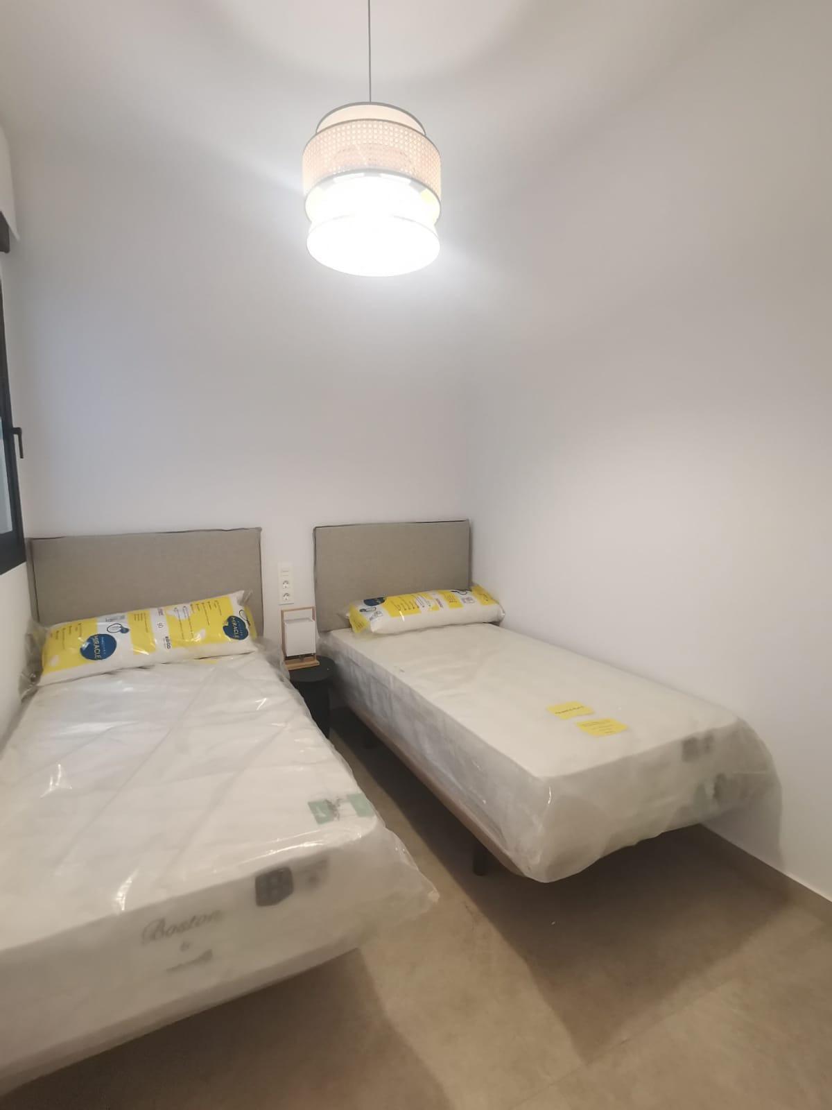 Small twin bedroom with two single beds in plastic, beige headboards, and a shared nightstand beneath a hanging ceiling light.