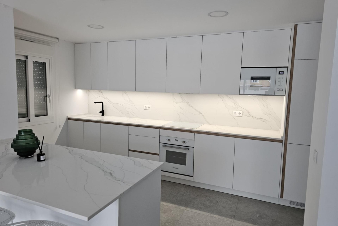 Modern white kitchen with marble countertops, grey cabinets, and built-in oven/microwave. Black faucet over sink, window with blind on left.