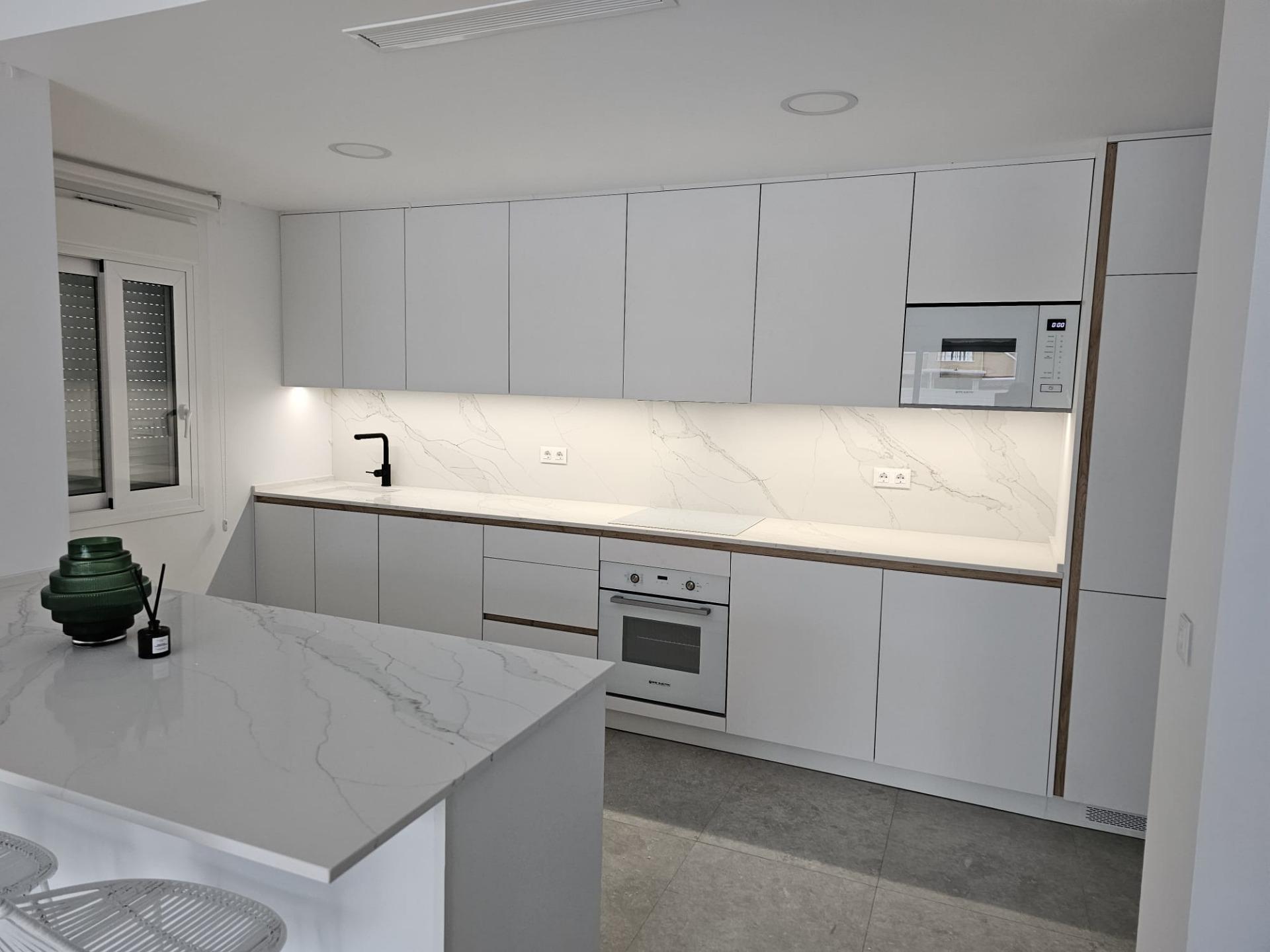 Modern white kitchen with marble countertops, grey cabinets, and built-in oven/microwave. Black faucet over sink, window with blind on left.