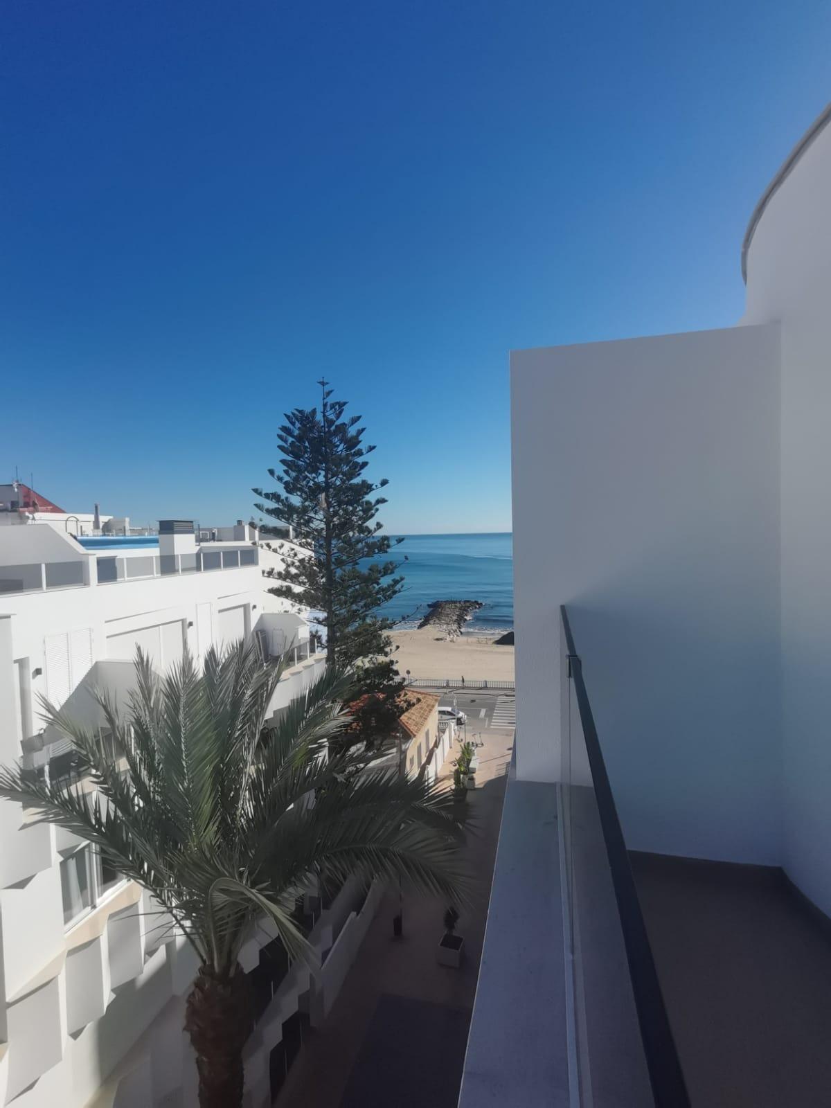 View from a balcony: white modern buildings along a palm-lined street with the blue sea and clear sky in the distance, near a sandy beach.