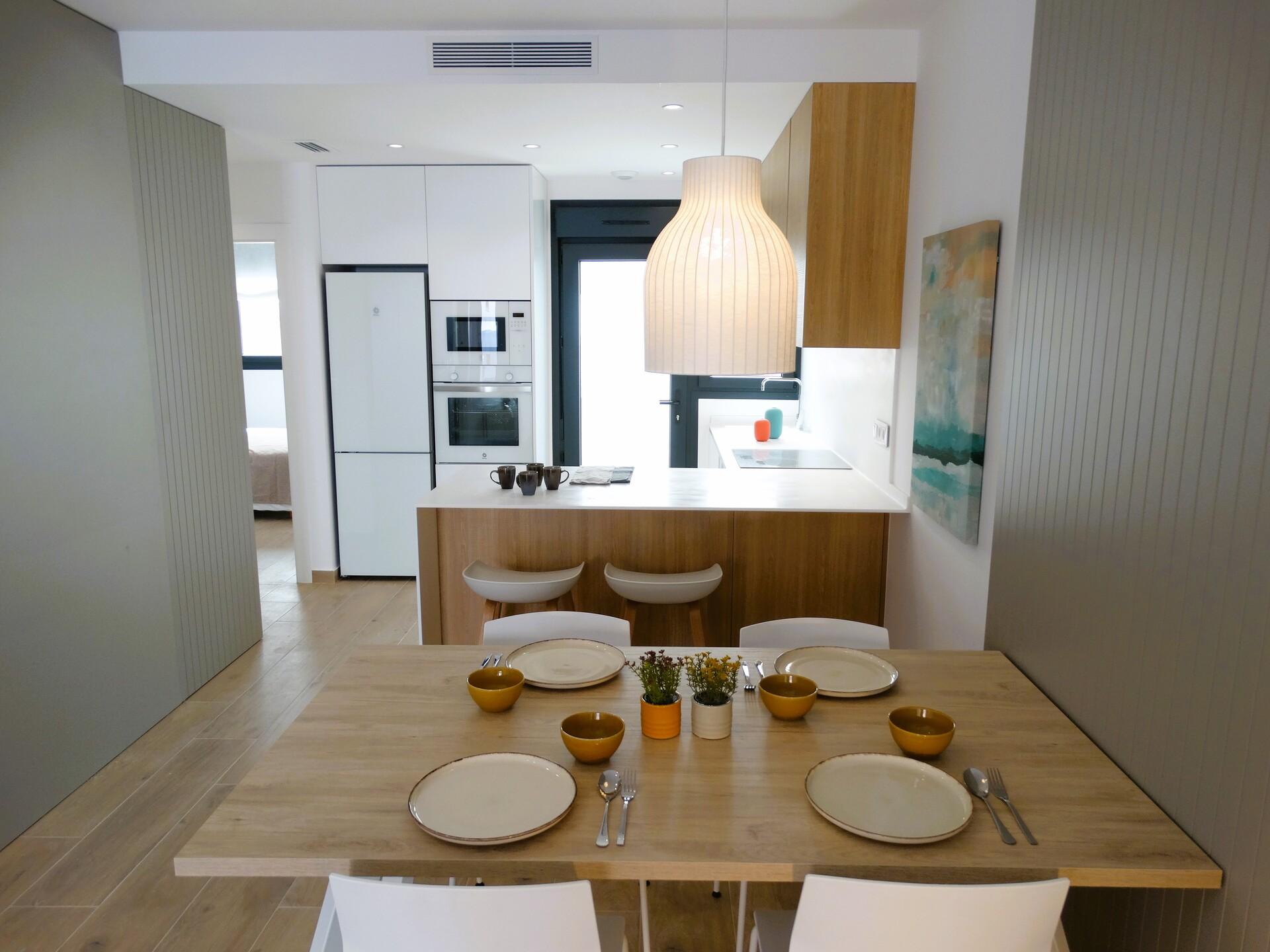 Modern kitchen and dining area with a wooden island, pendant lamp, and a table set for a meal with white plates and yellow cups on a light wood floor.