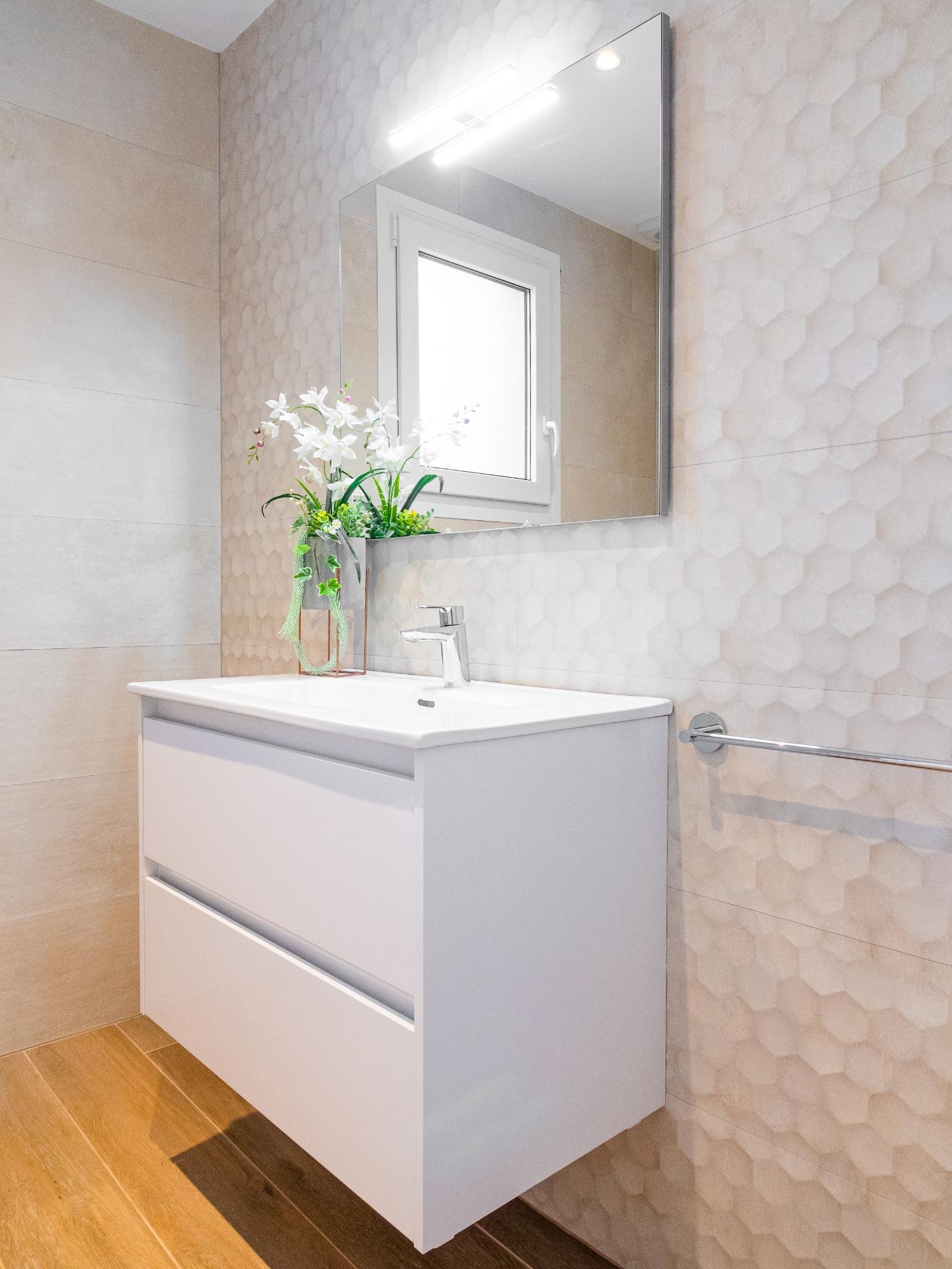 Modern bathroom vanity with a white cabinet, rectangular sink, and chrome faucet, reflected in a large wall mirror above; beige hexagonal-tile wall and a potted orchid plant on the sink.