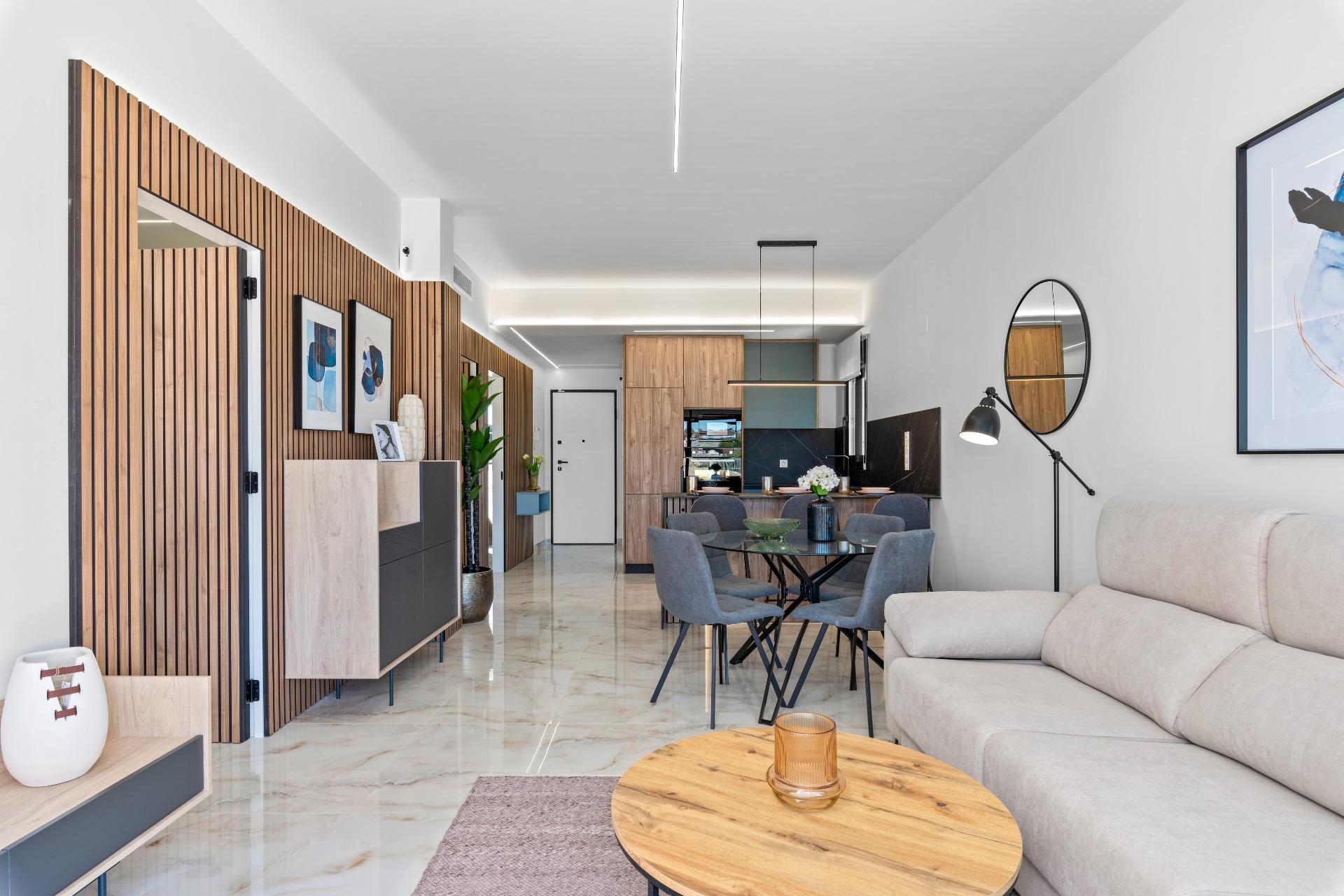 Open-plan living and dining area with a gray sofa, round wooden coffee table, marble floors, and a wood-slatted wall facing a modern kitchen.