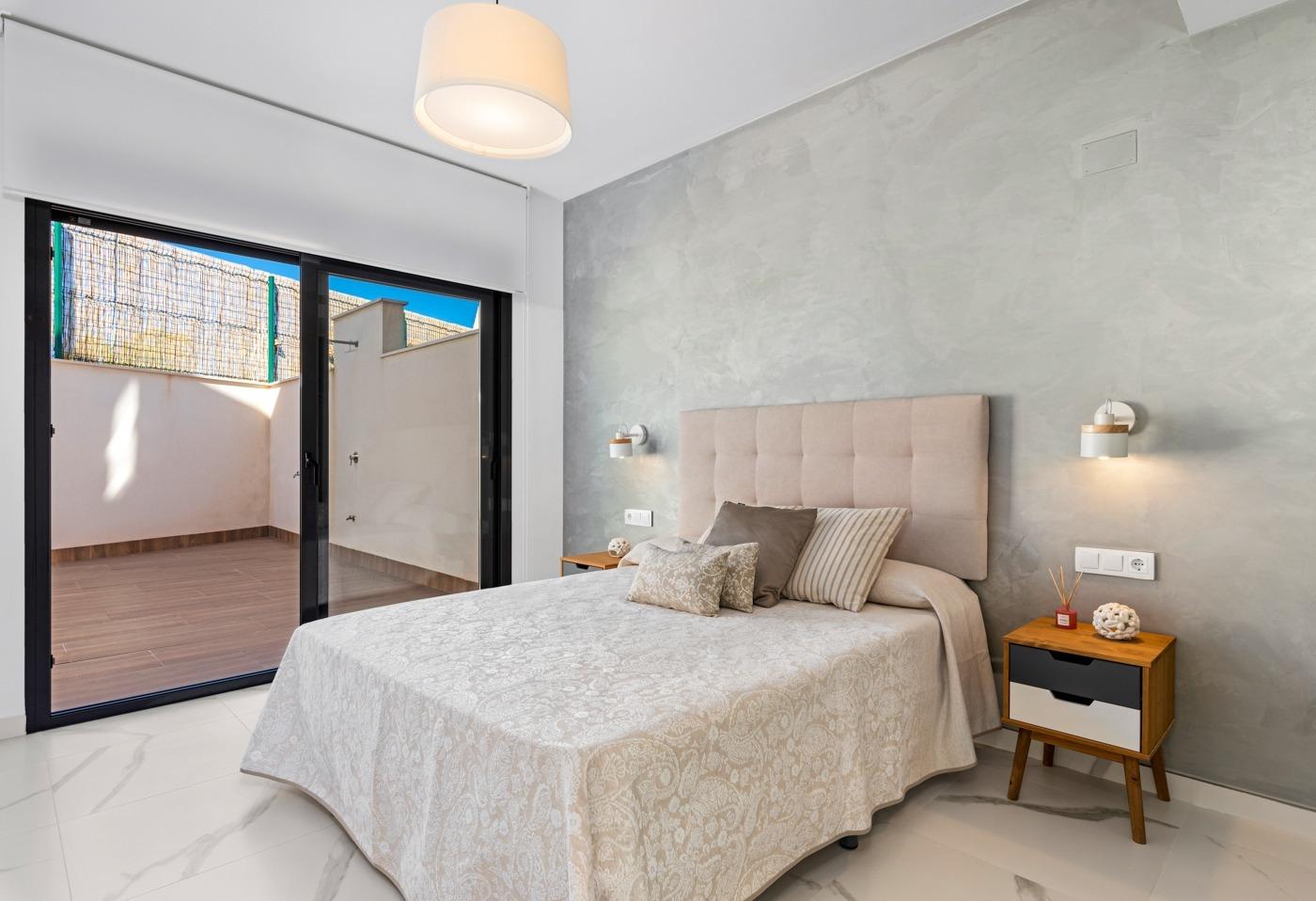 Bedroom with a tufted beige headboard, patterned bedspread, and two wooden nightstands; sliding glass doors open to a small terrace. The room has a light gray textured wall and white marble floor.