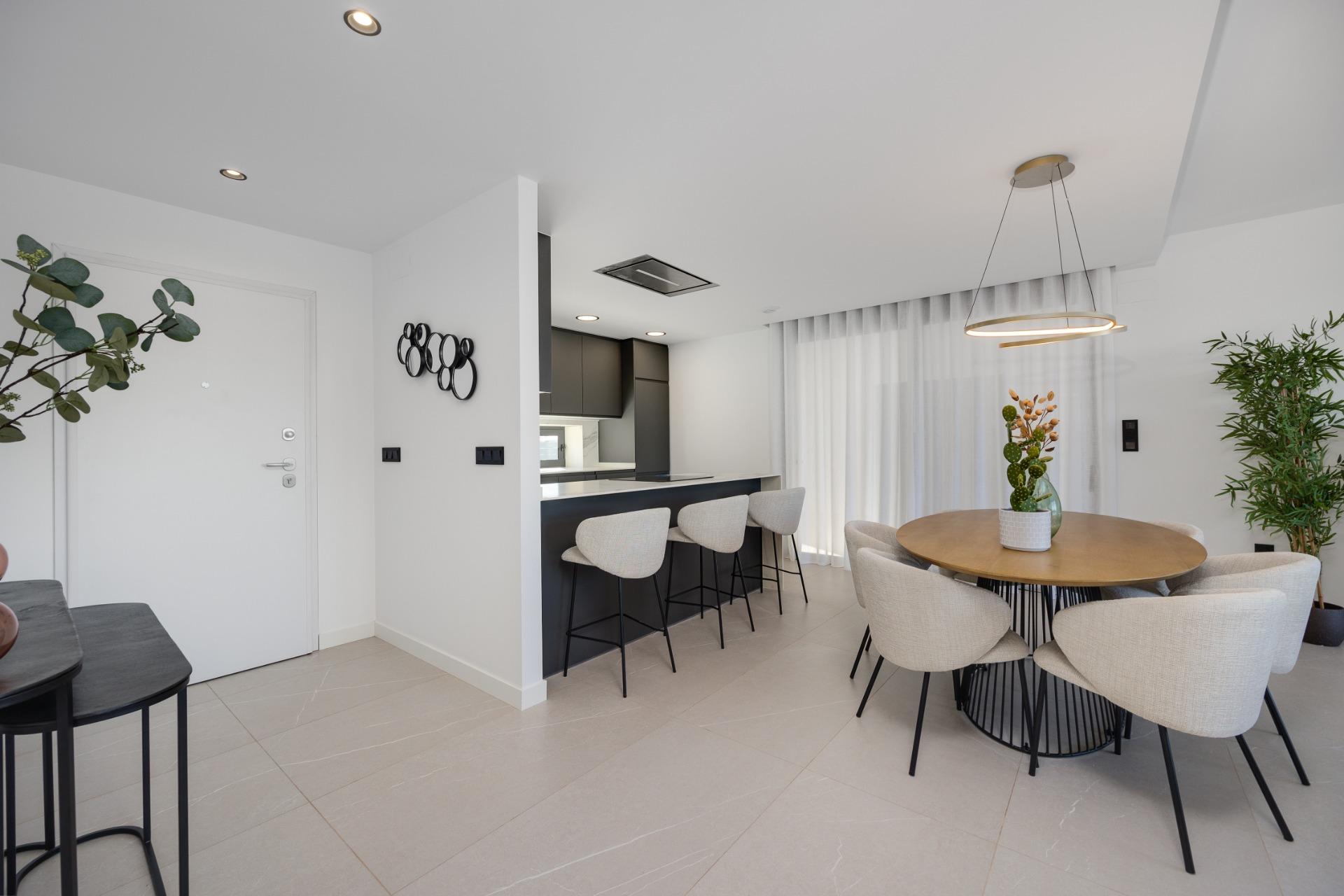 Open-plan dining and kitchen area with round wood table, beige upholstered chairs, and a ring pendant light; white walls and large windows with sheer curtains.
