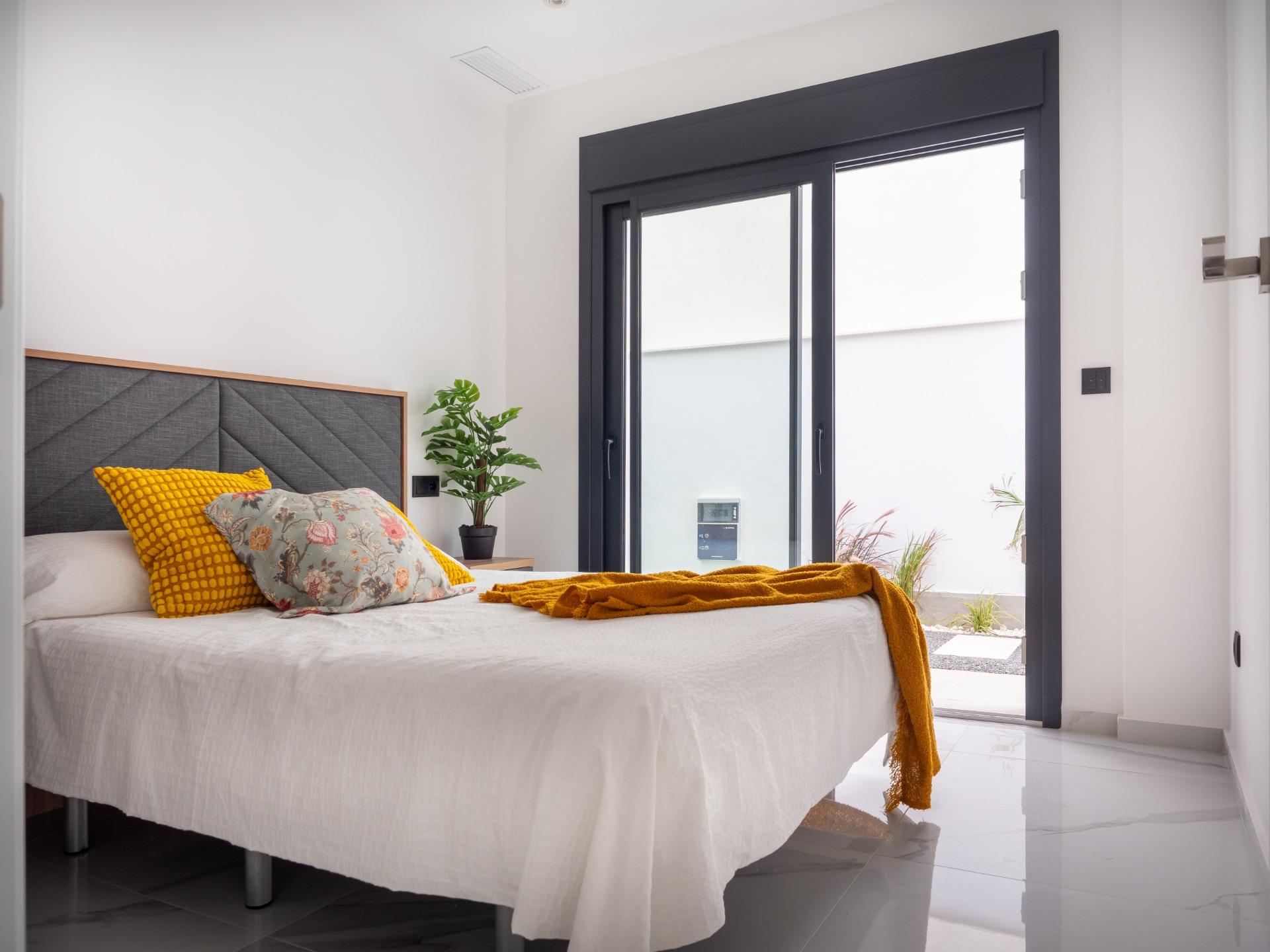 Bright modern bedroom with white bedding, yellow throw and floral pillows on a gray upholstered headboard. Sliding glass doors open to outside with a plant nearby.
