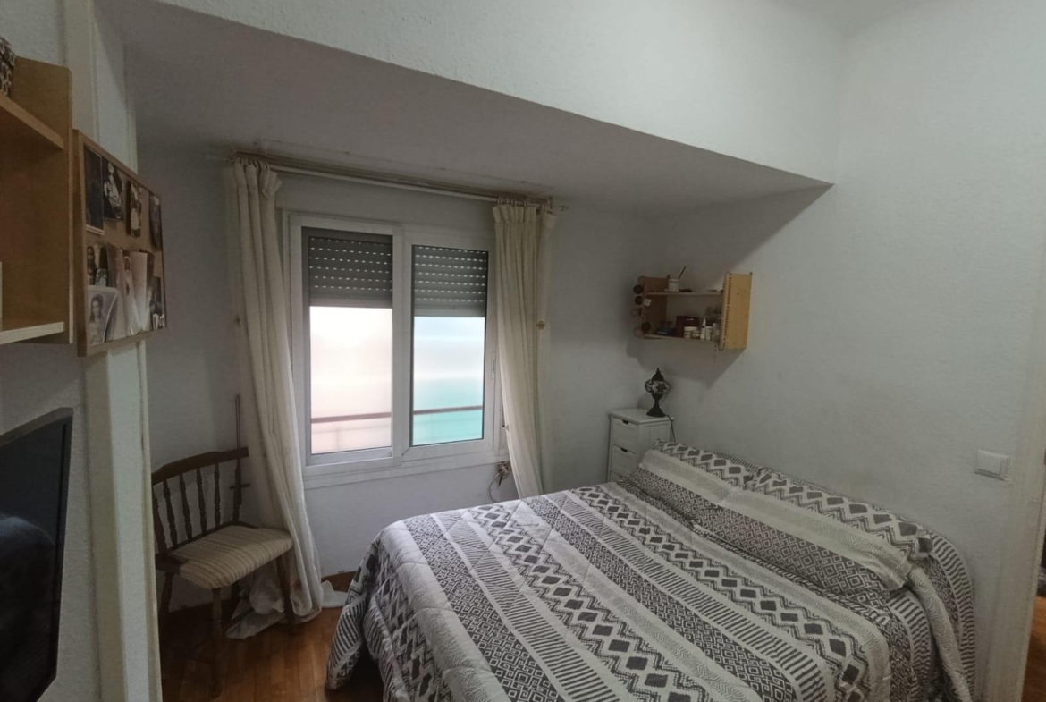 Cozy bedroom with a double bed in the foreground, a window with blinds and white curtains, and a small dresser against the wall near a decorative shelf.