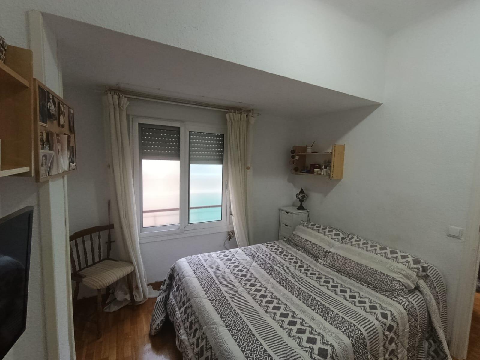 Cozy bedroom with a double bed in the foreground, a window with blinds and white curtains, and a small dresser against the wall near a decorative shelf.