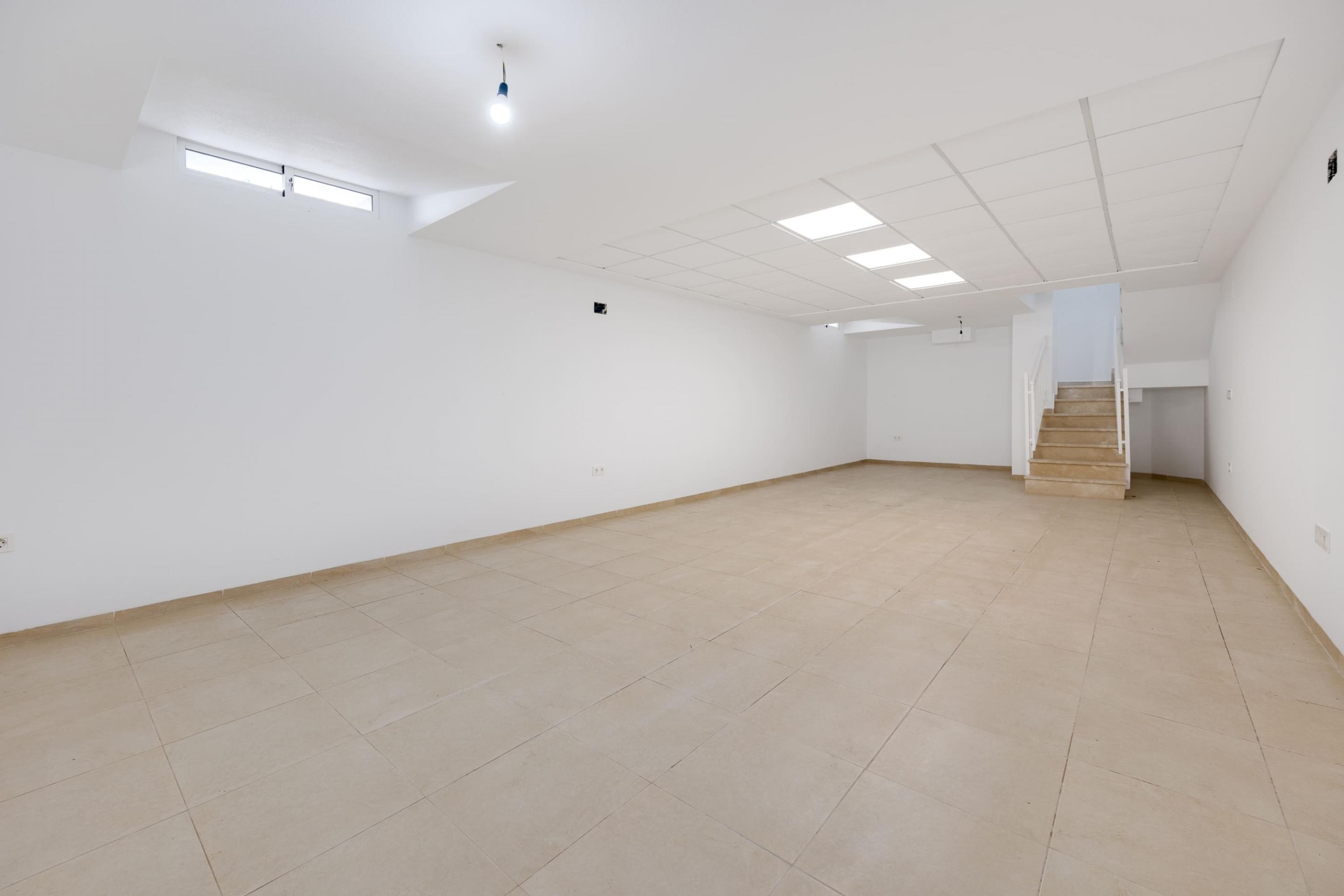 Wide empty room with white walls and beige tile floor, bright ceiling lights, and a stairway on the right.