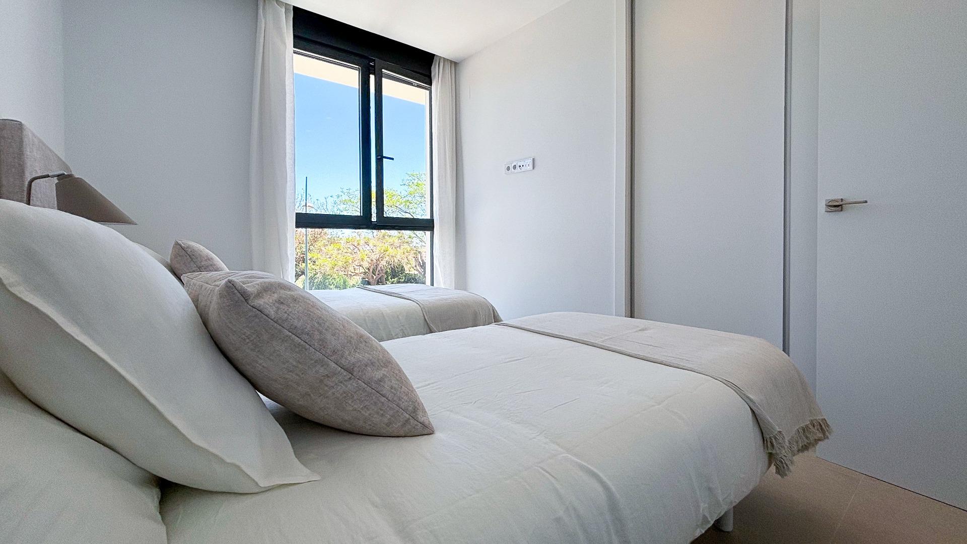 Two neatly made twin beds in a bright minimalist bedroom with white bedding and gray pillows, next to a window with a view of trees outside.