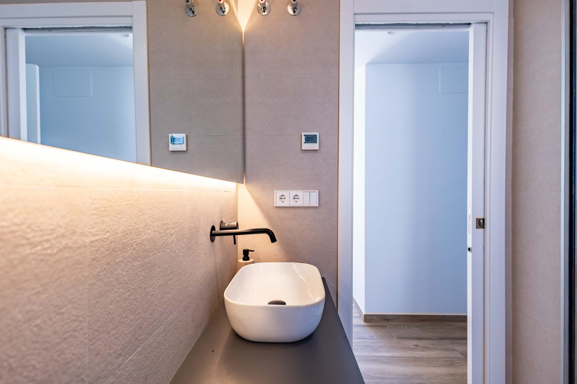 Modern bathroom with a white vessel sink on a dark countertop, black wall-mounted faucet, soap dispenser, and a doorway to an adjacent room separated by glass doors.