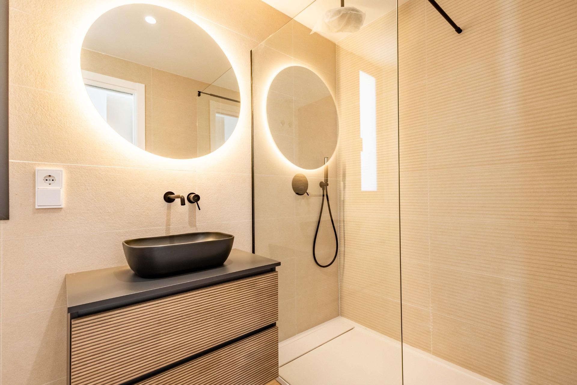 Modern bathroom featuring a round backlit mirror, black vessel sink on a ribbed wooden vanity, and a glass shower enclosure.