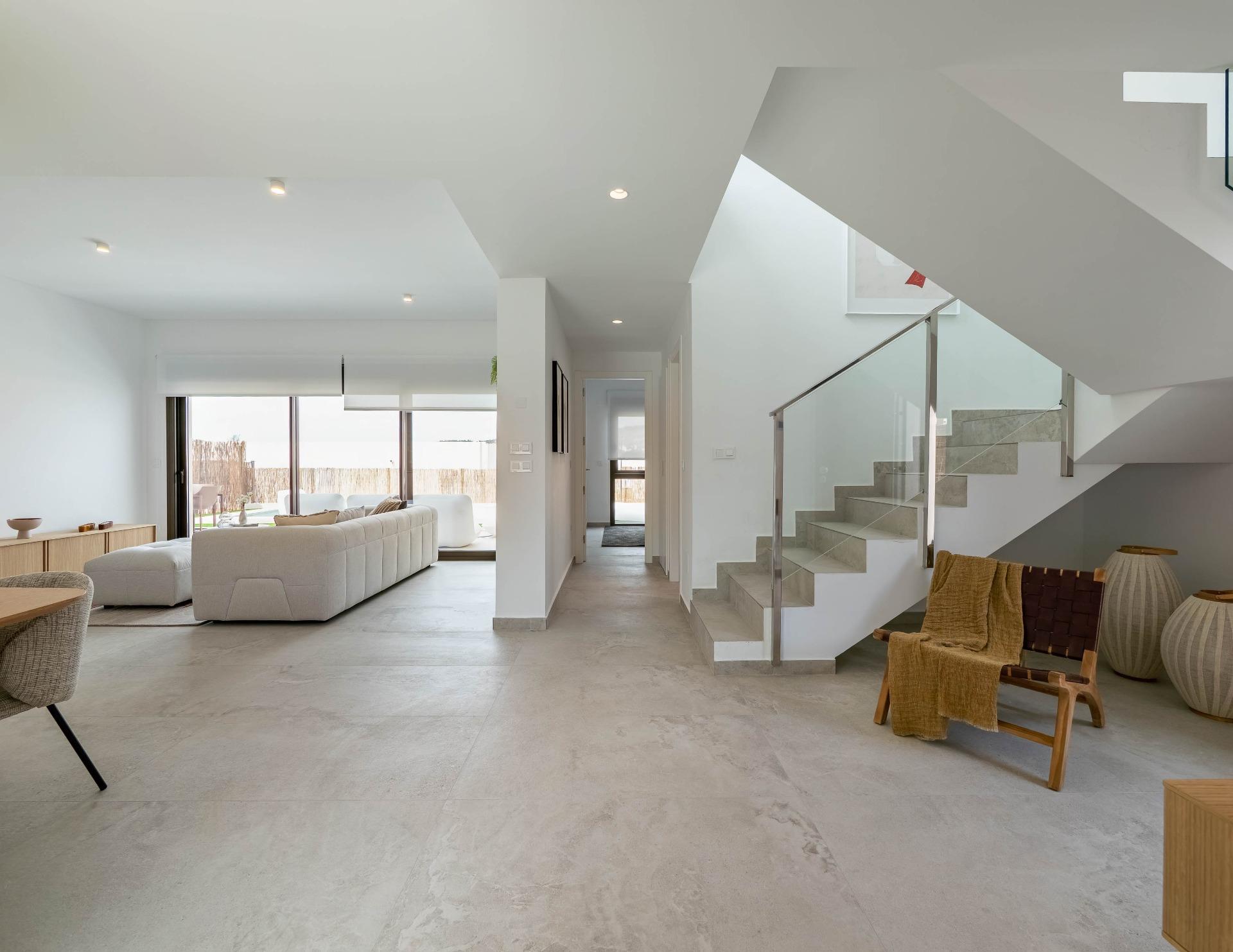 Bright open-concept living area with pale gray flooring, white walls, a large gray sofa, and a glass-railed staircase on the right.