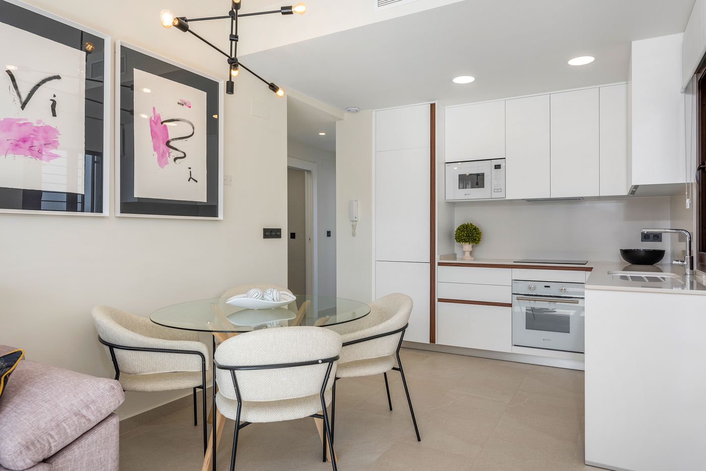 Open-concept dining area with a glass round table, four cream chairs, and abstract black-and-pink wall art on a light wall.