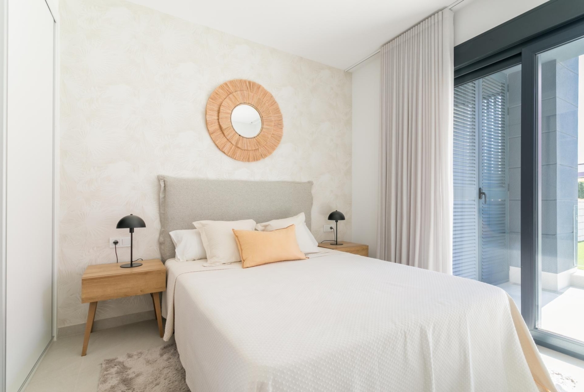 Bright hotel bedroom with white bedding, beige headboard, two wooden nightstands, black lamps, a woven circular mirror, and balcony doors.