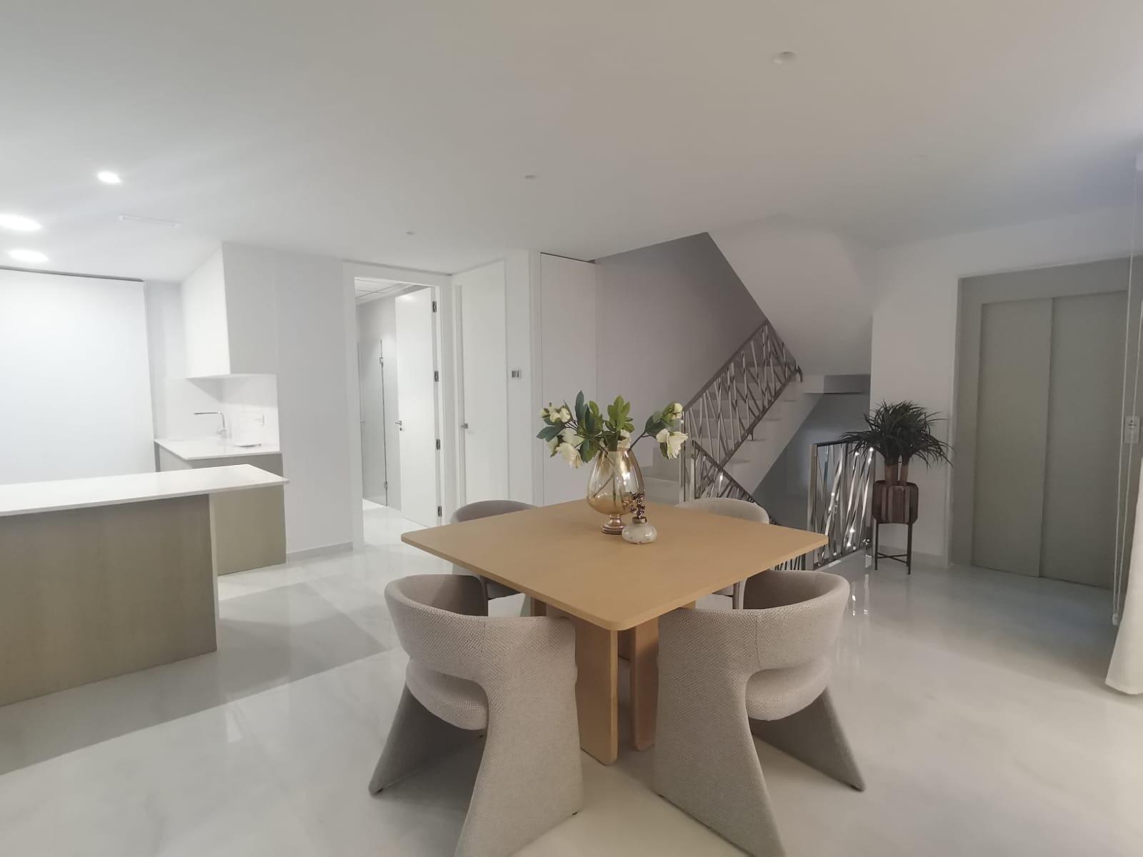 Open-plan dining area with a square wooden table and six beige upholstered chairs, a vase of white flowers in the center, and a metal-railed staircase on the right.