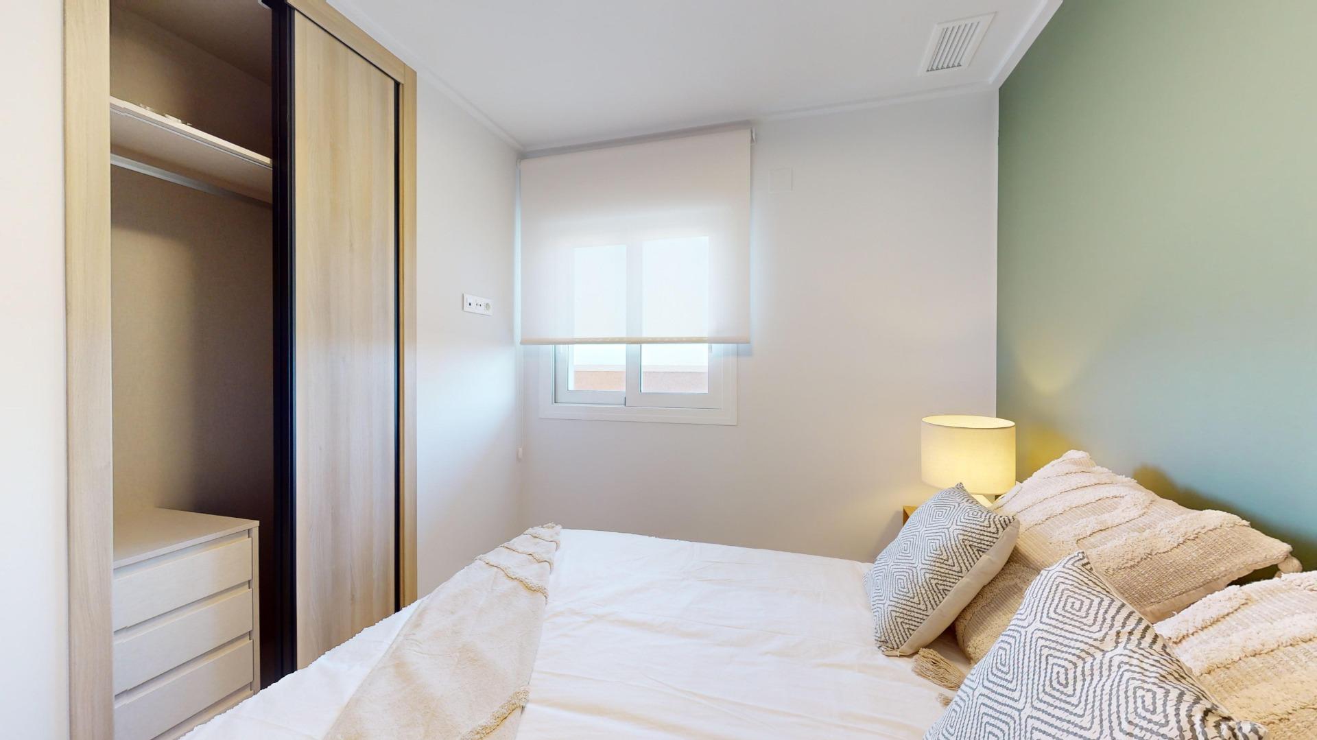 Small bedroom with a double bed, beige bedding and patterned pillows, a light wood wardrobe on the left, and a green accent wall on the right.