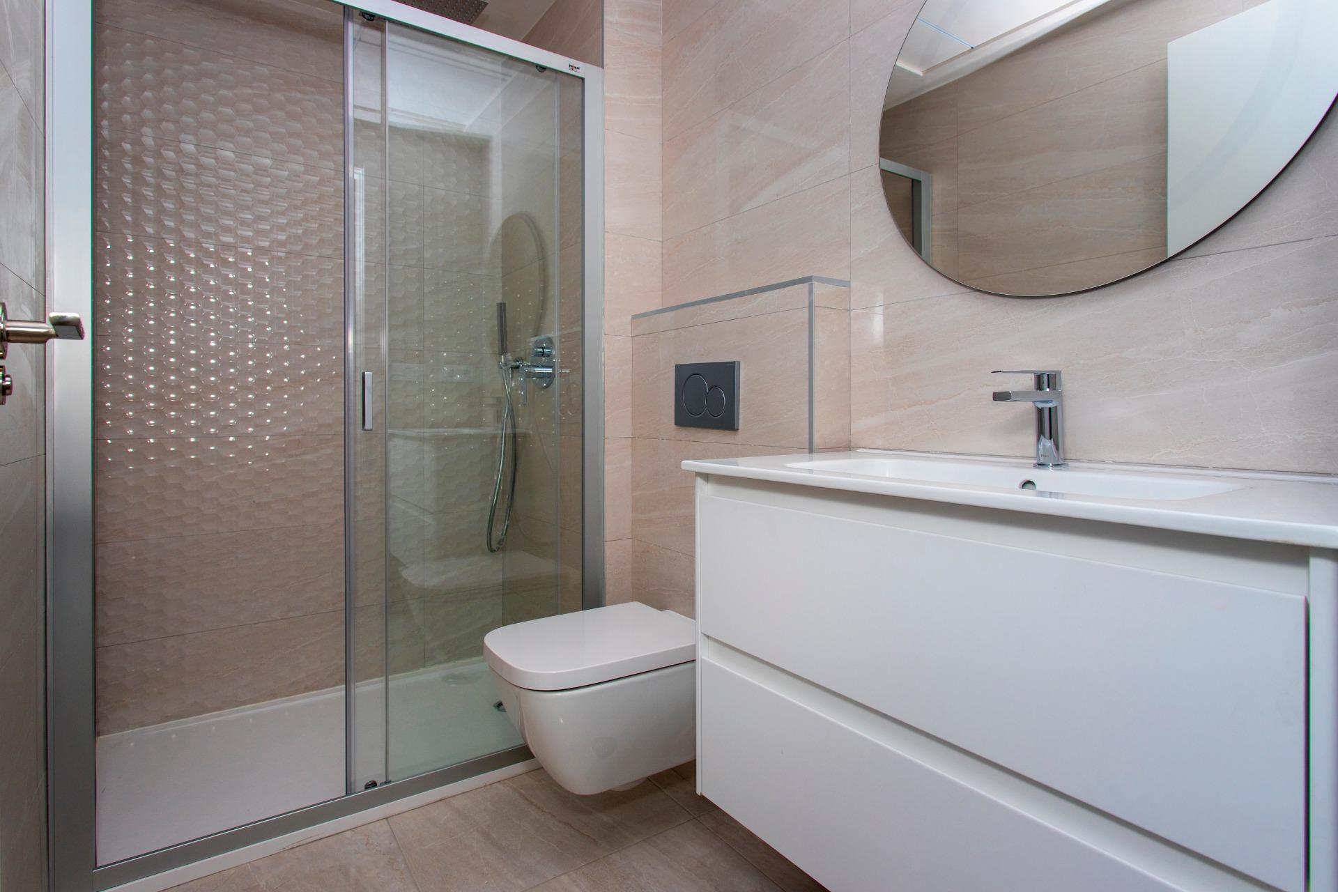 Modern bathroom with a glass shower enclosure, wall-mounted toilet, and white vanity beneath a round mirror.