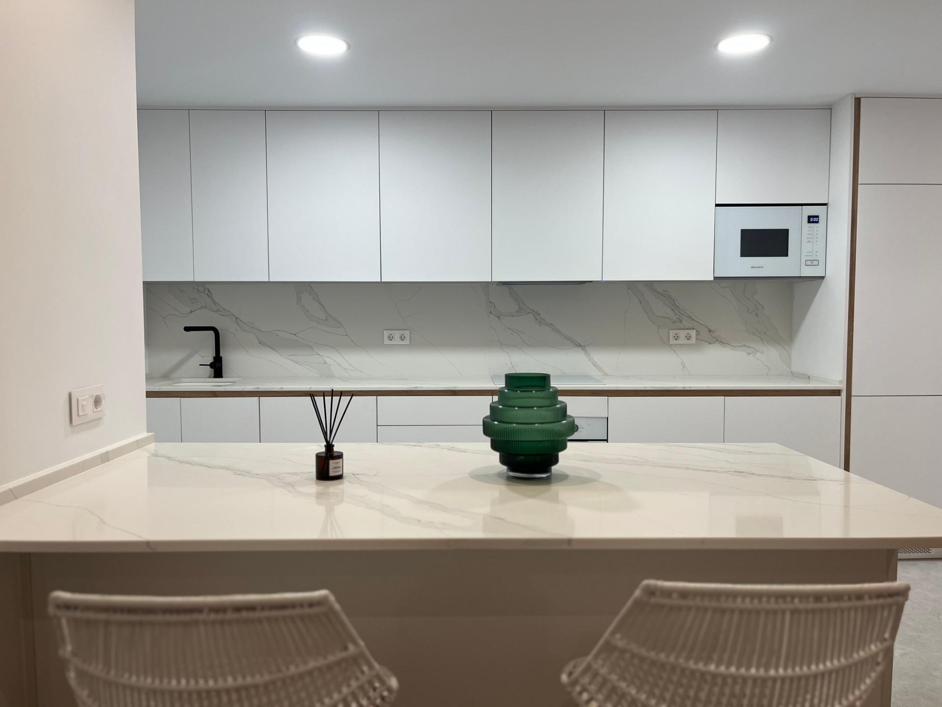 Modern kitchen with white cabinets and marble countertop; island in foreground, green glass tiered centerpiece, and a reed diffuser in a small bottle on the counter