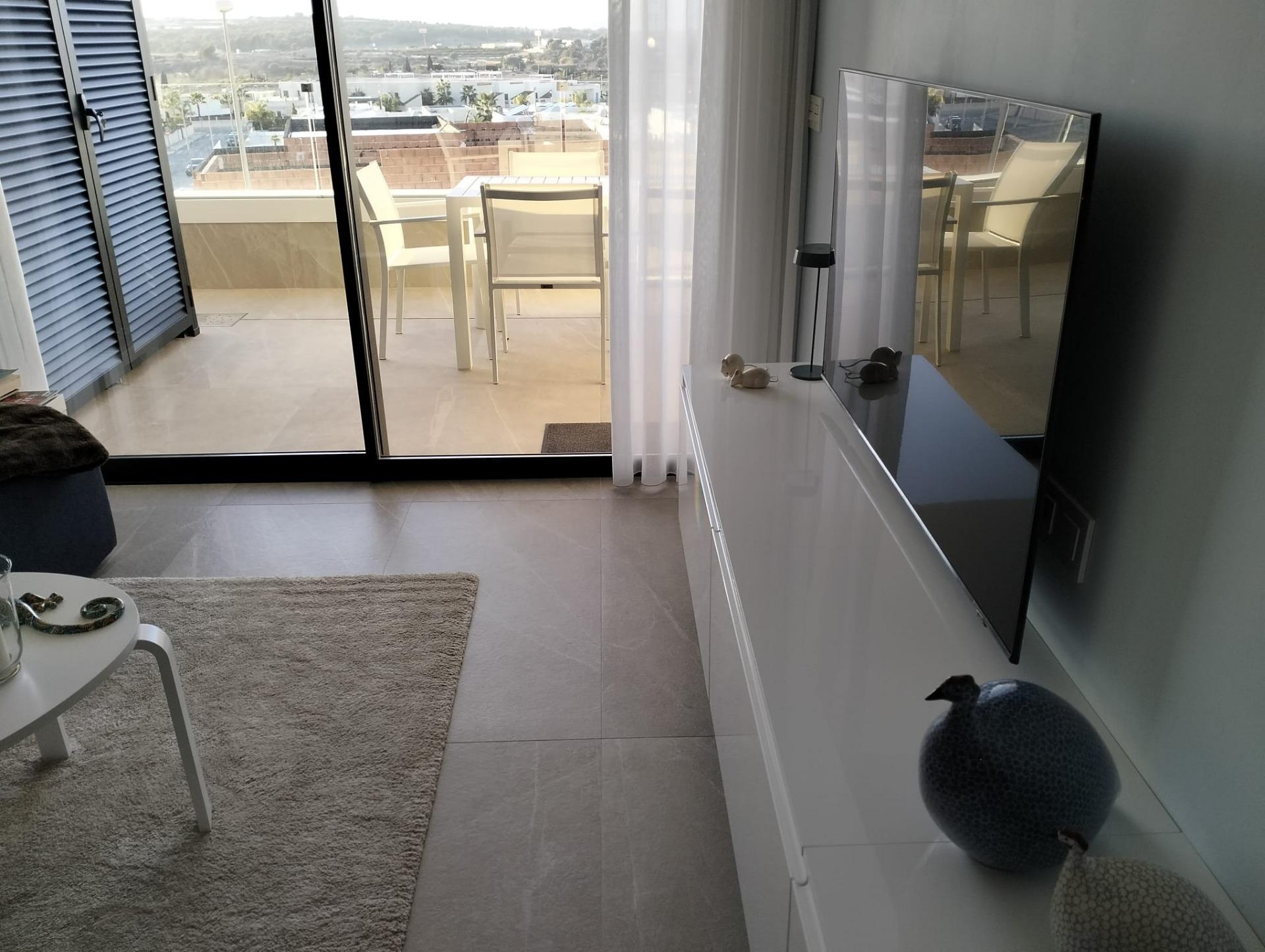 Living room with a balcony view: glass doors, white furniture, and a TV on a white cabinet; outdoor table and chairs on the balcony beyond.