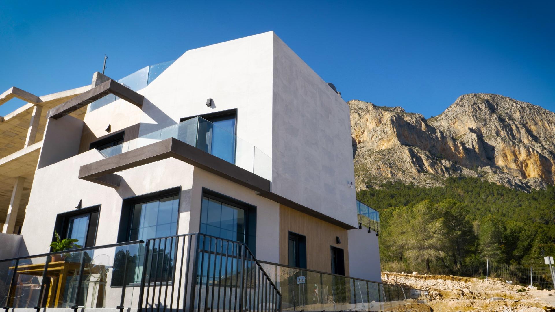 Modern white house with glass balconies against a rocky mountain backdrop and clear blue sky.