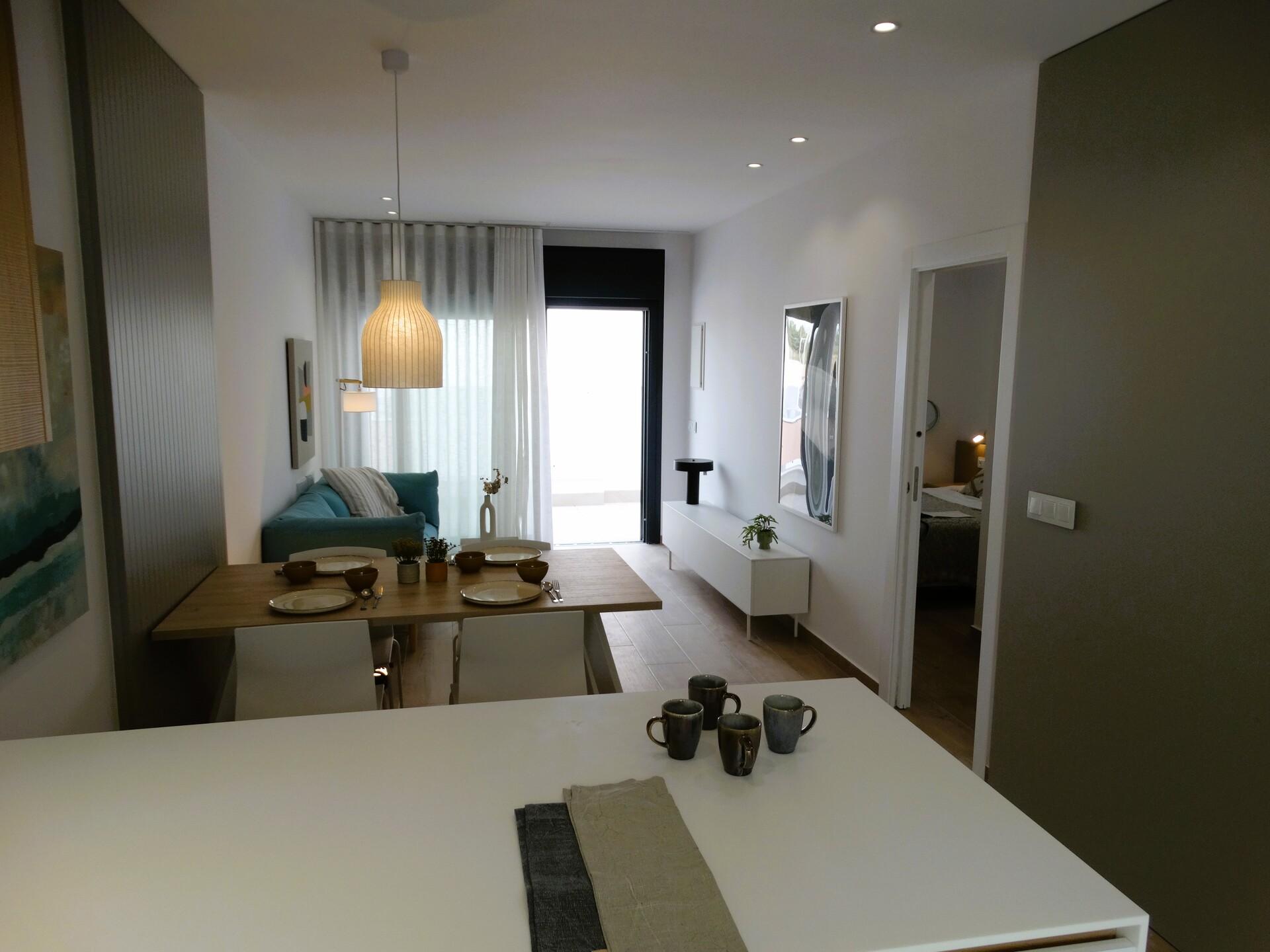 Open-plan living and dining area with a wooden table set for six, teal sofa, hanging pendant light, and large glass door with sheer curtains.