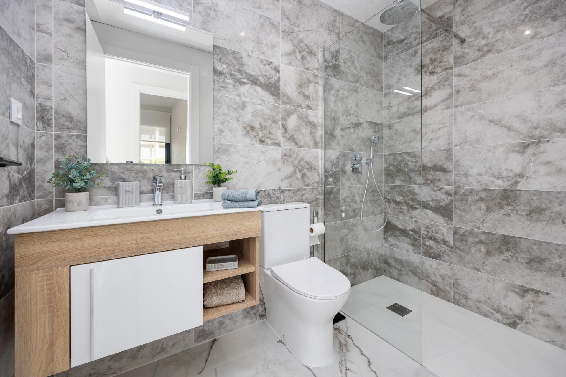 Modern bathroom with marble walls, wooden vanity, and glass shower enclosure; sink area with plants, towels, and toiletries visible.